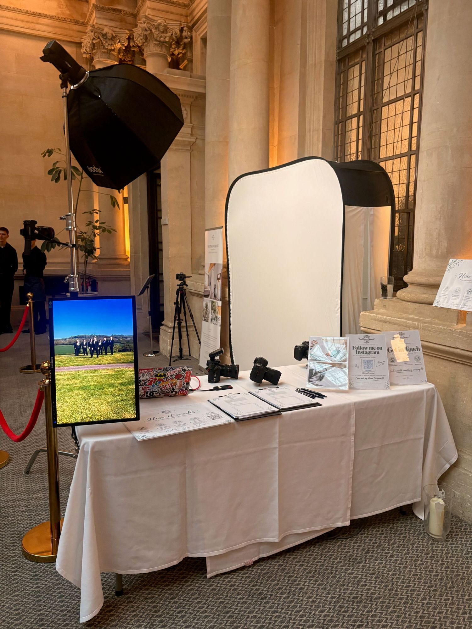 Professional photo booth setup with lighting equipment, white backdrop, camera, and display monitor at an indoor event venue.