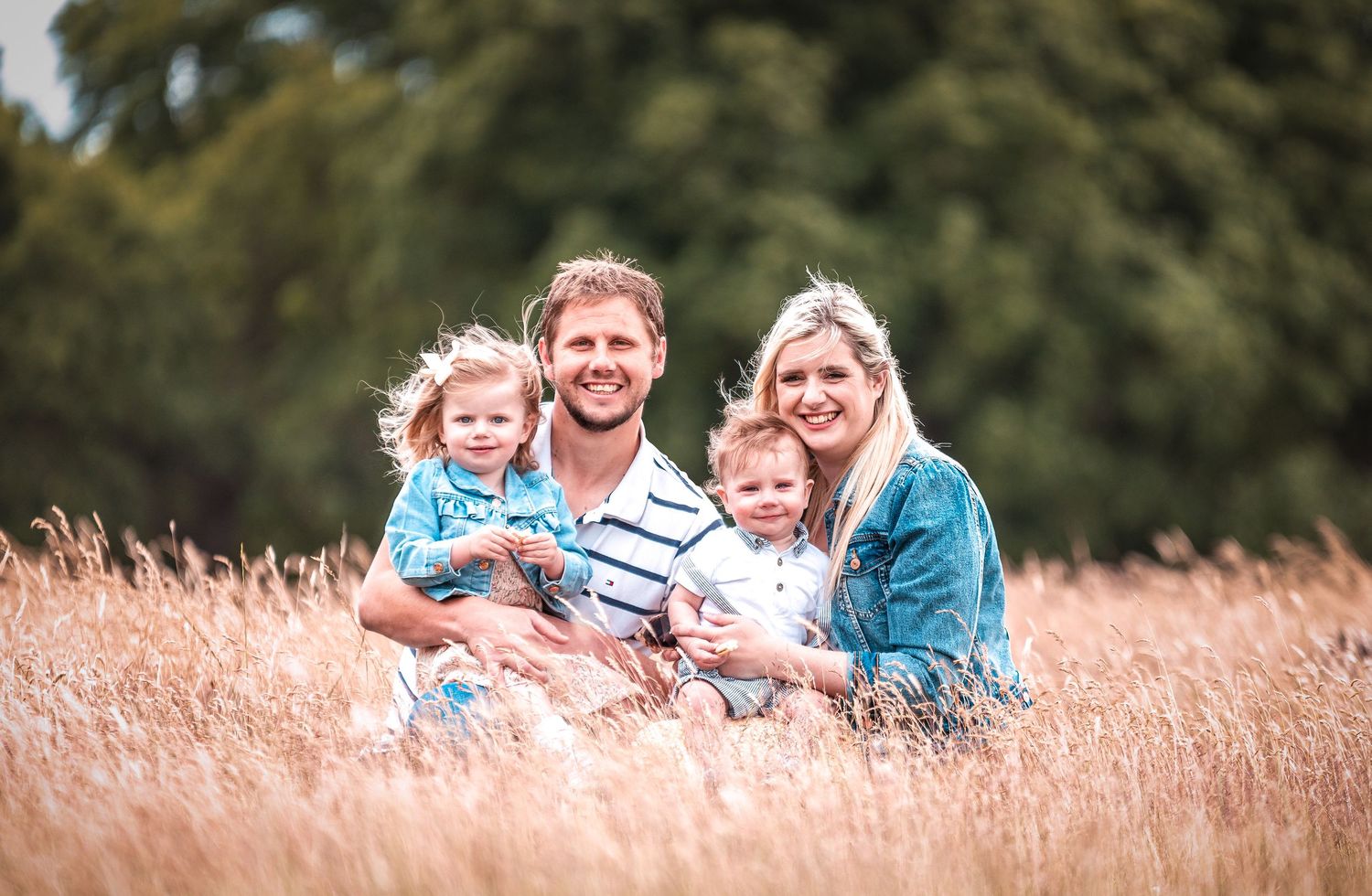 A happy family sits together in a sunlit field of tall wheat grass during golden hour.
