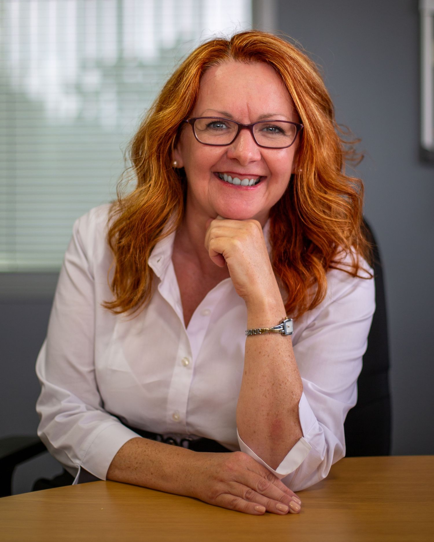Professional wearing glasses and white button-down shirt smiles while resting at desk in office setting.