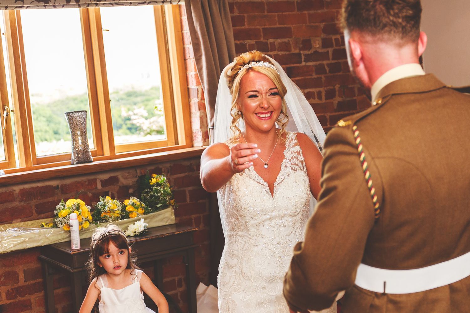 A joyful wedding ceremony moment in front of a brick wall with large windows, featuring military dress uniform and bridal attire.