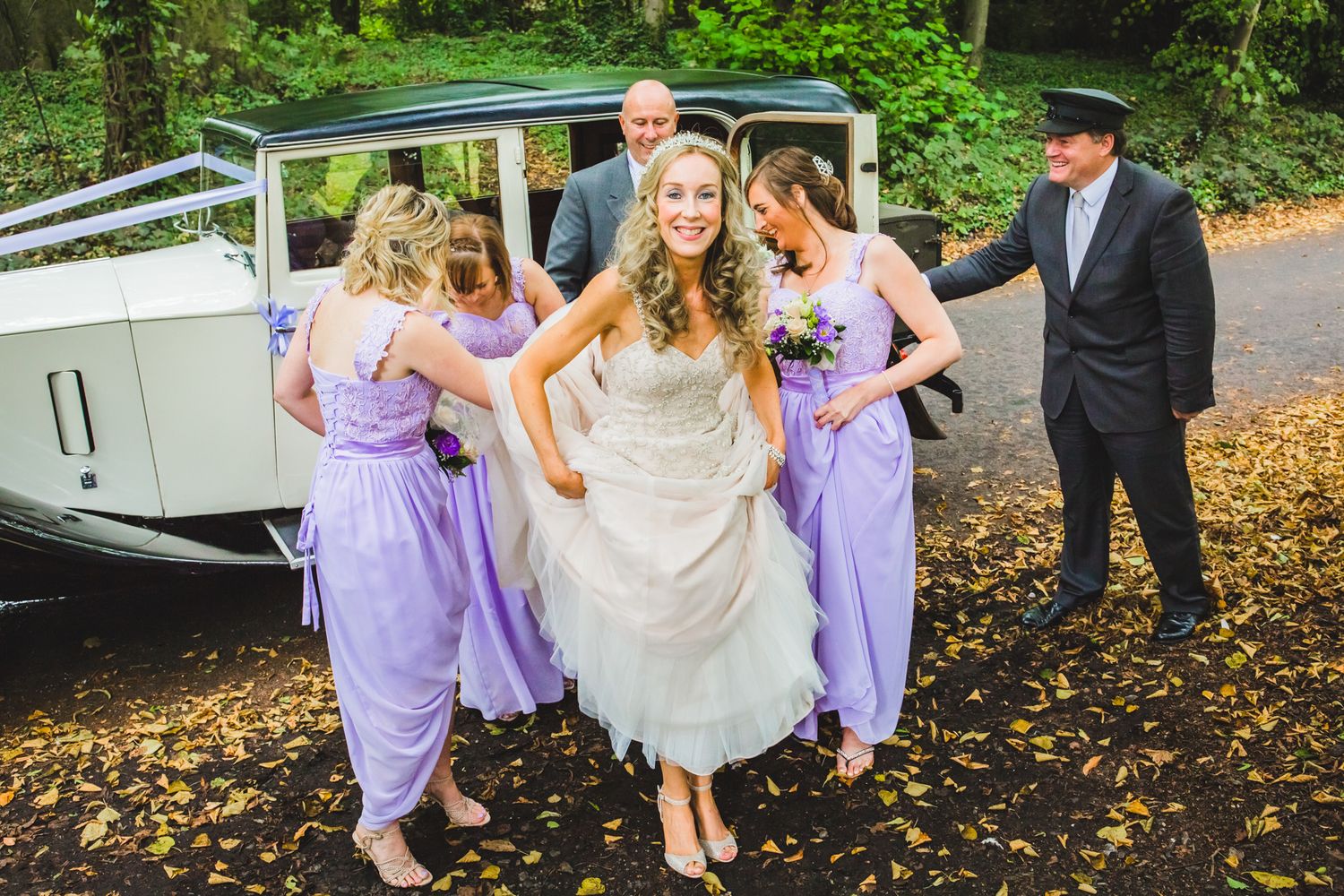 Wedding party in light purple bridesmaid dresses exit vintage white car on autumn day with fallen leaves.