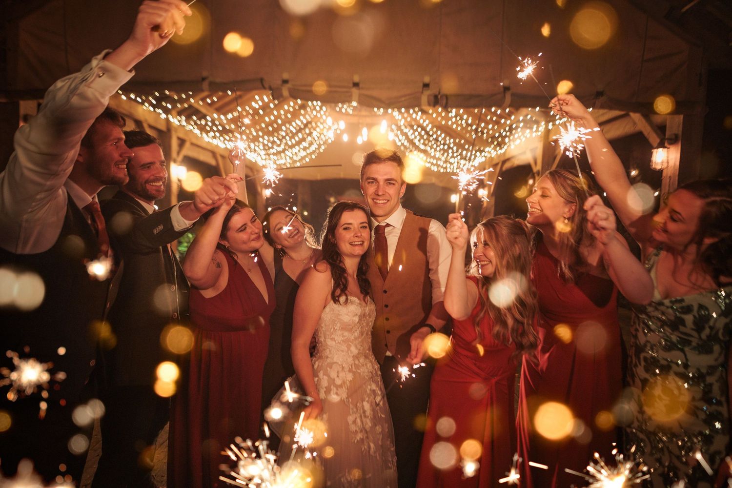 Wedding party dances and celebrates under twinkling lights and sparklers at an evening reception.