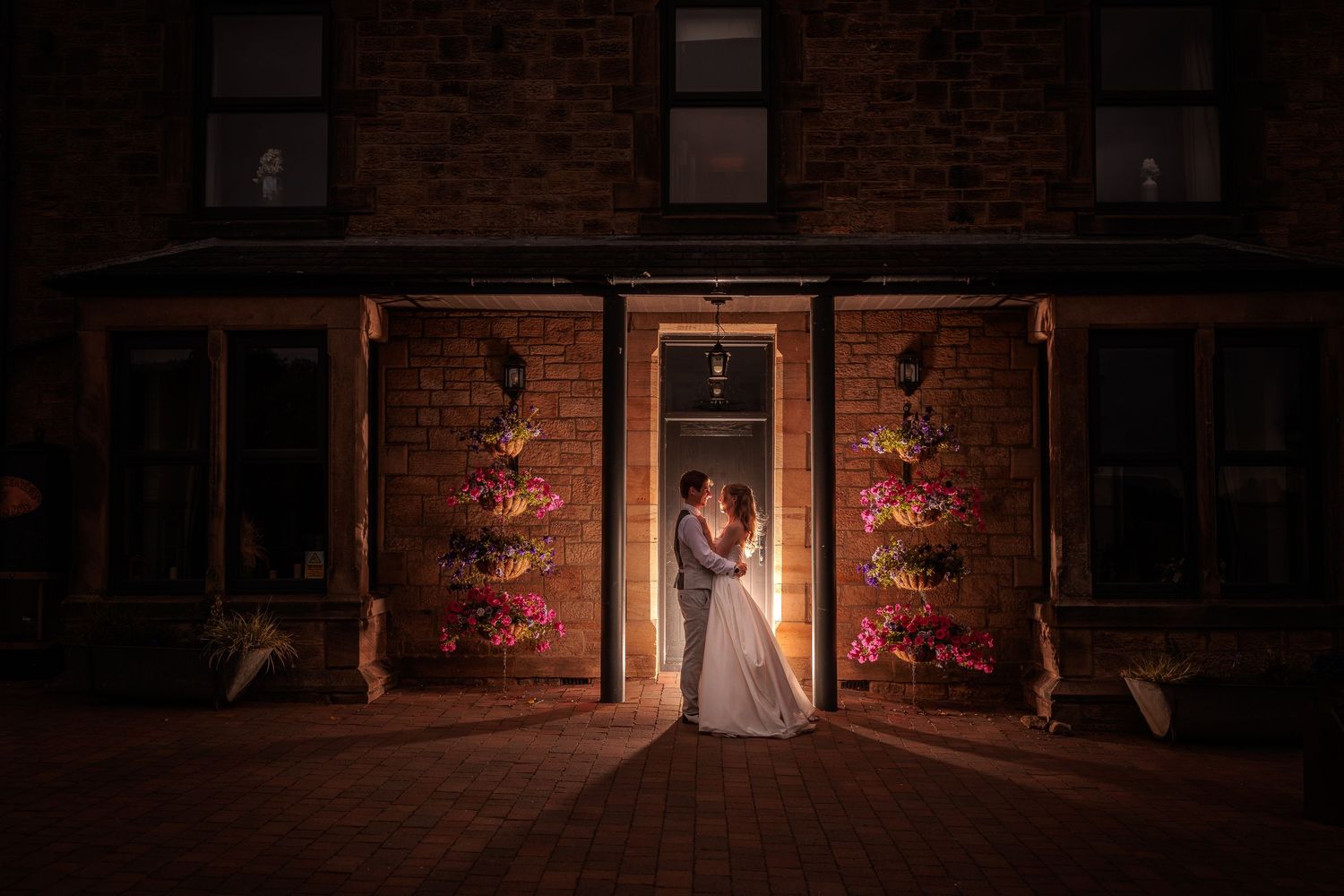 A dramatic night wedding photo with uplighting on a brick wall and soft spotlights creating a romantic atmosphere.