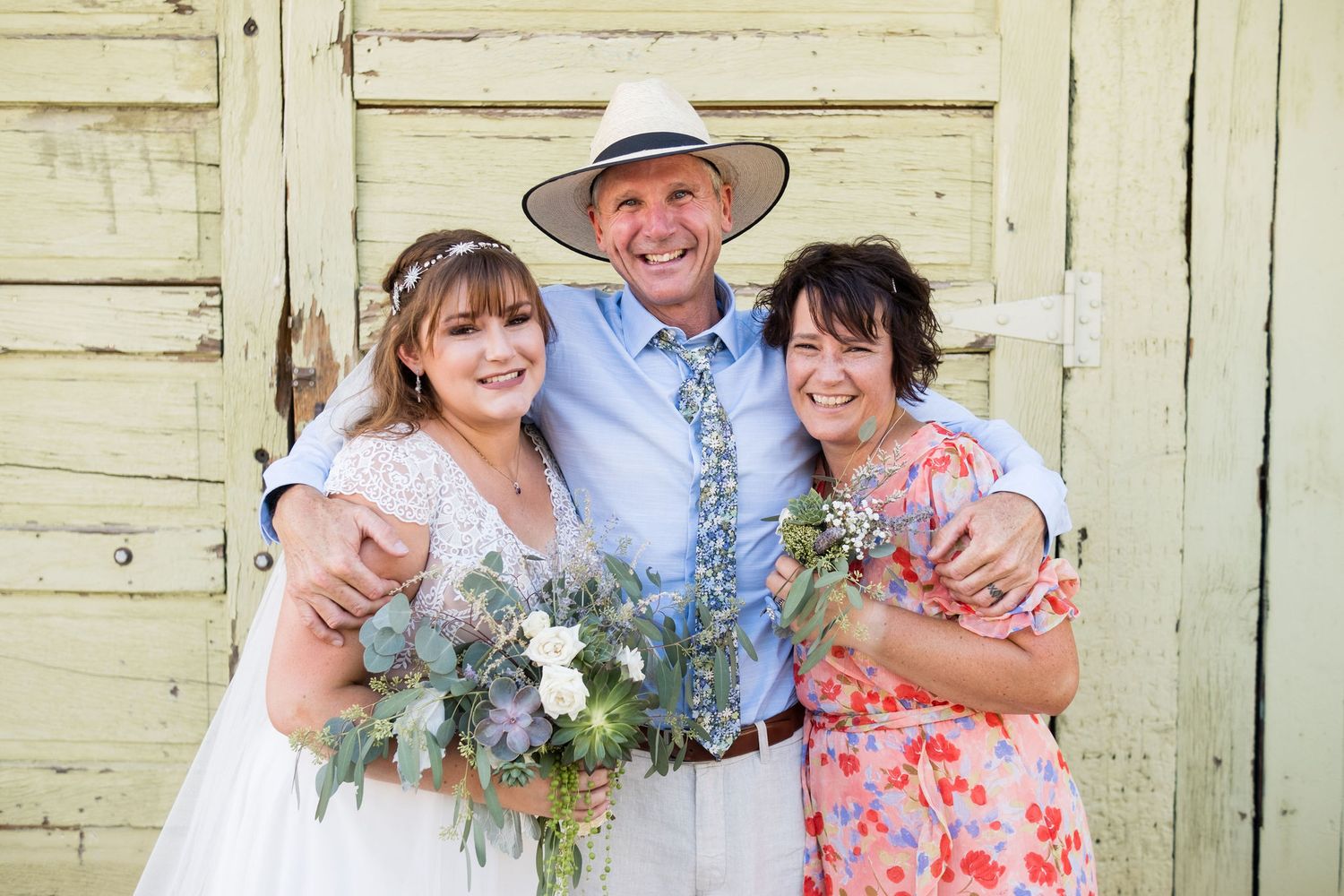 Bride in white dress holding bouquet poses with two guests in front of rustic yellow barn doors.