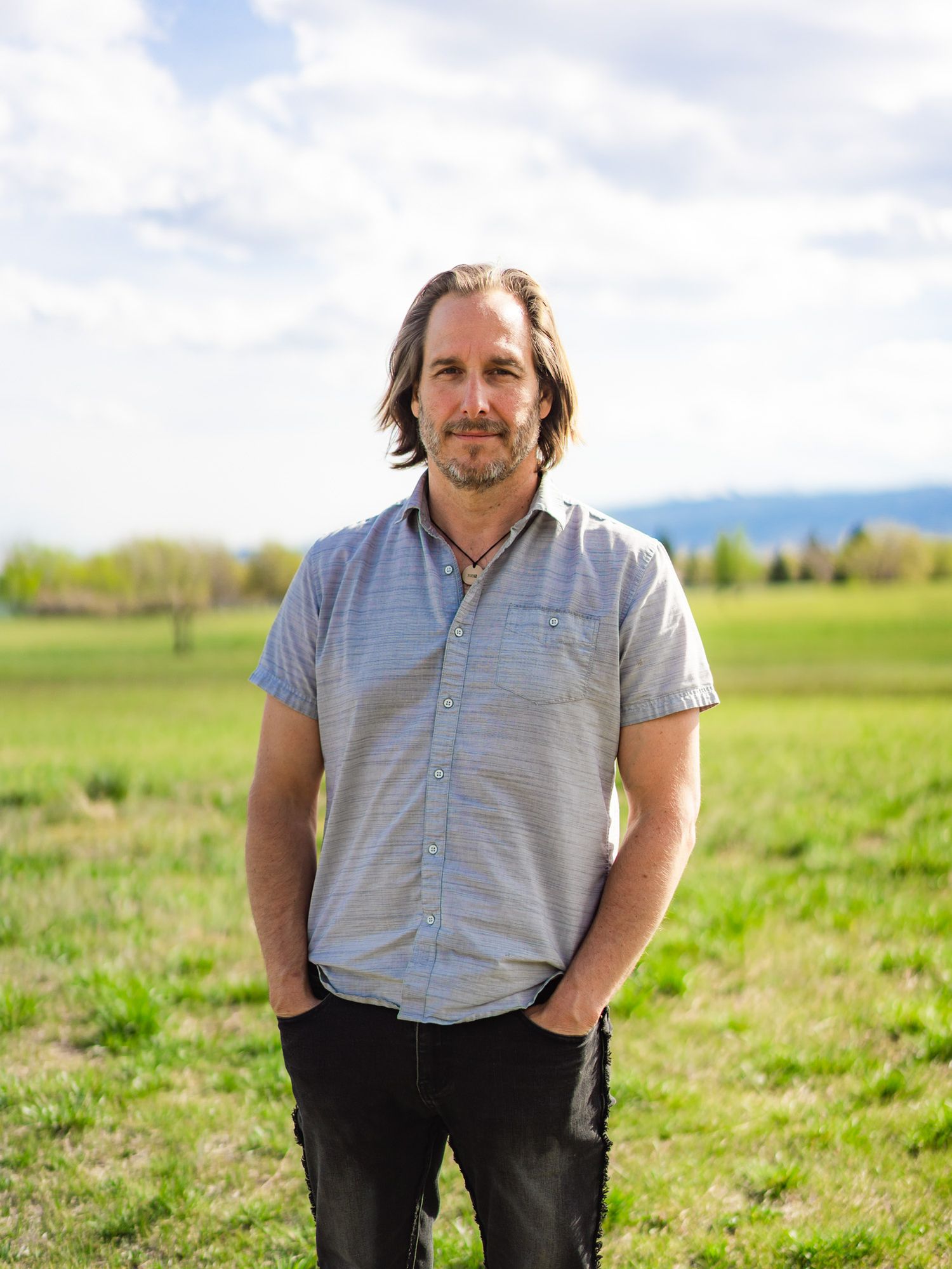 Man with shoulder-length hair stands in a green field with mountains and cloudy sky in the background.