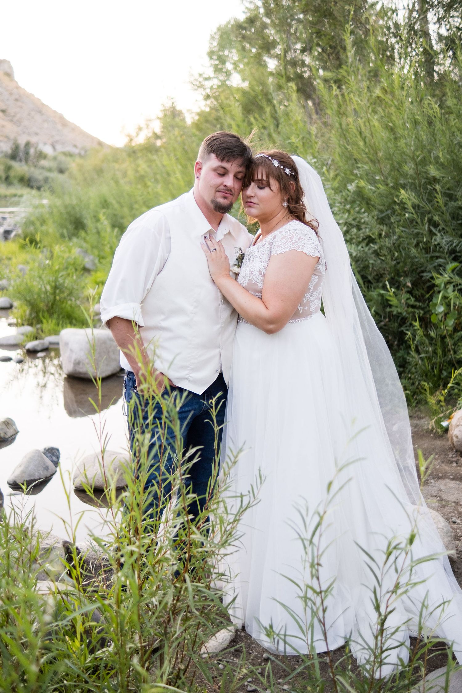 Bride and groom pose romantically near a rocky stream surrounded by lush greenery on their wedding day.