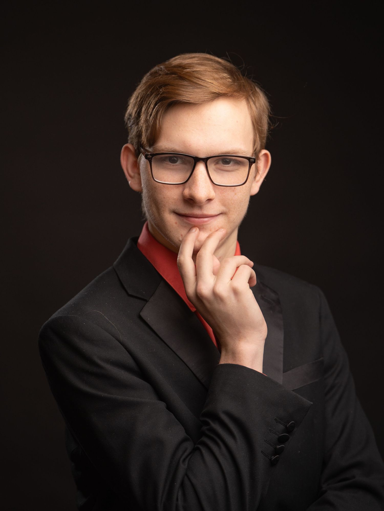 Professional studio portrait of an actor in black suit jacket and red shirt wearing glasses against dark background.
