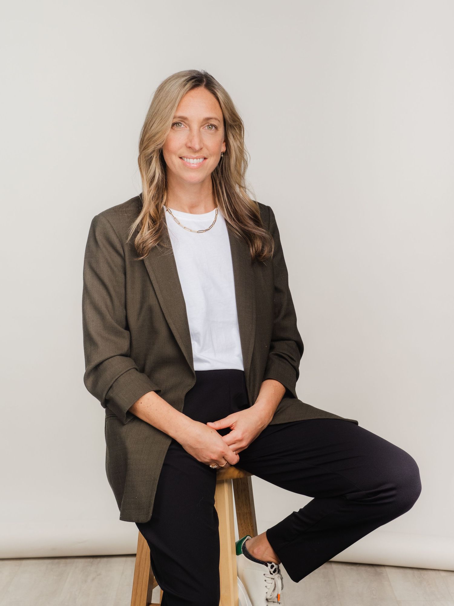 Professional portrait of someone in a brown blazer, white t-shirt and black pants sitting casually on a wooden stool.