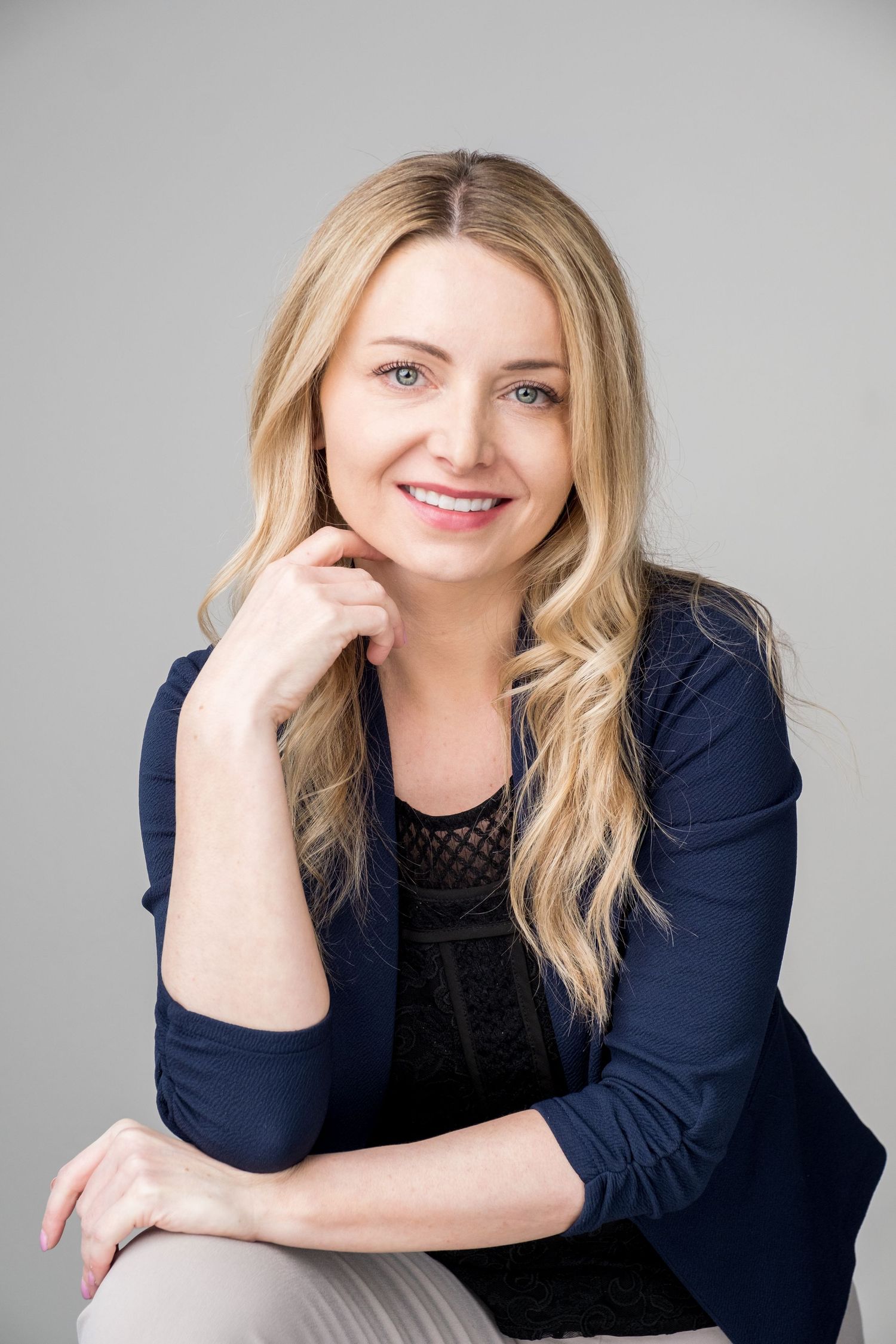Professional portrait of a young woman in a navy blazer and black top with blonde wavy hair against a gray background.