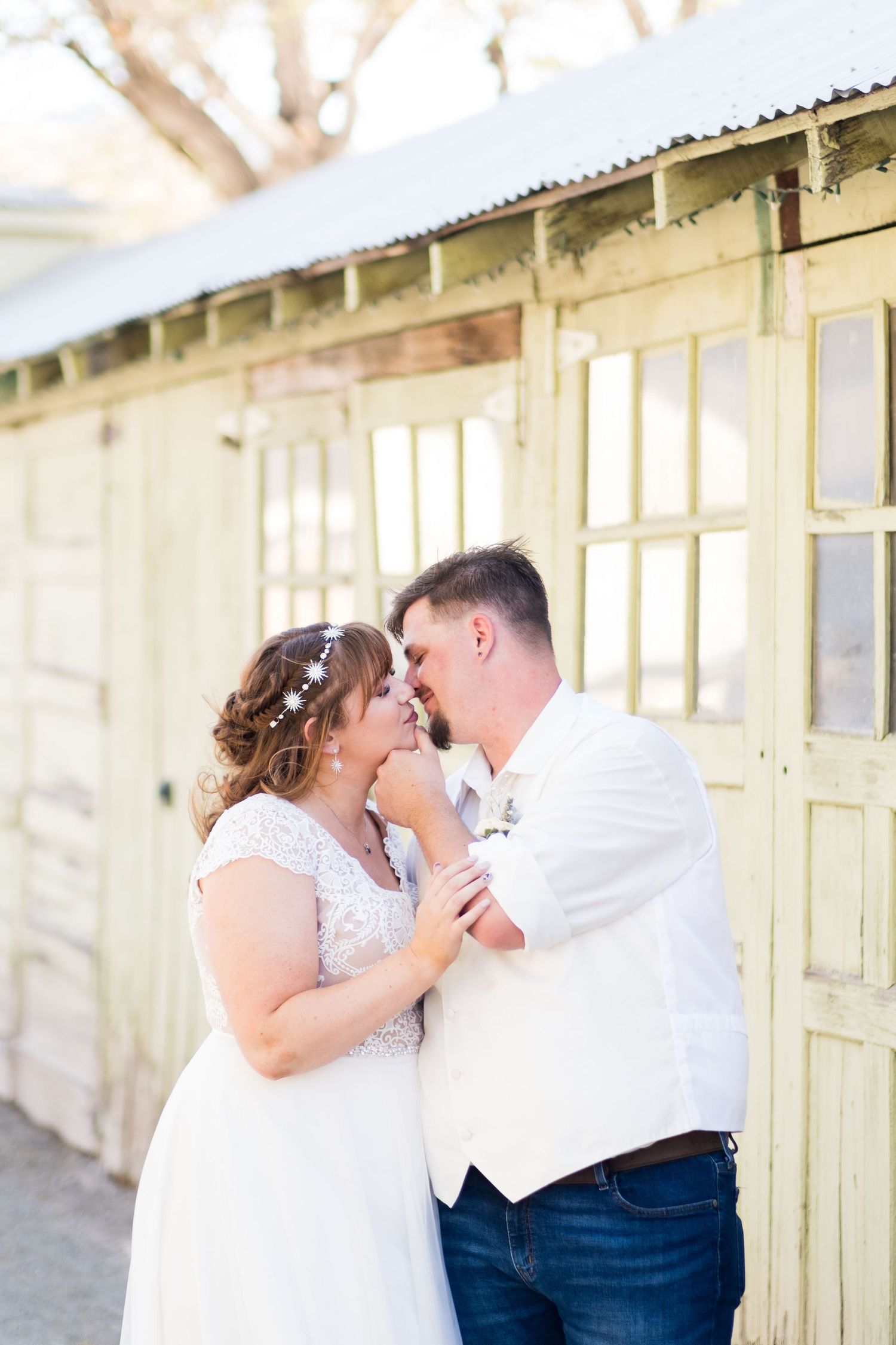 Bride and groom share a romantic moment, touching foreheads outside a rustic white wooden building on their wedding day.