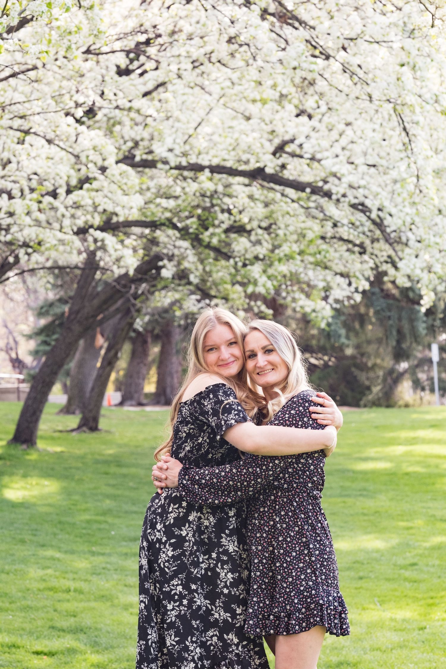 Mother and daughter in floral print dresses embrace under blooming cherry blossom trees in a spring garden.