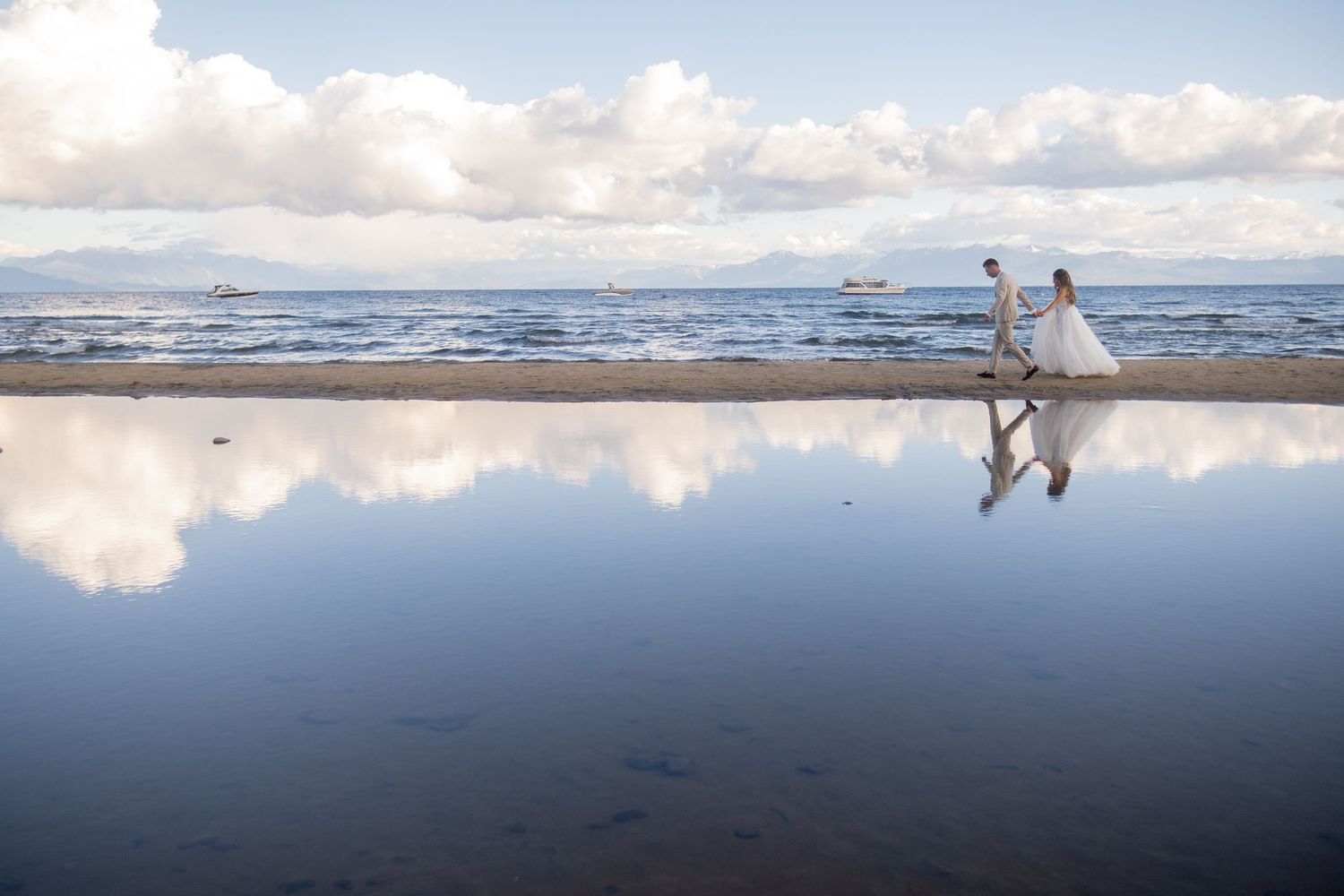 Bride and groom walk along a reflective beach shoreline with ocean and clouds mirrored in the wet sand.
