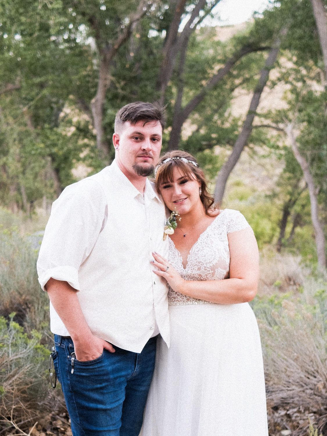 Bride and groom pose outdoors in rustic wedding attire, she wears a lace gown with floral crown, trees in background.