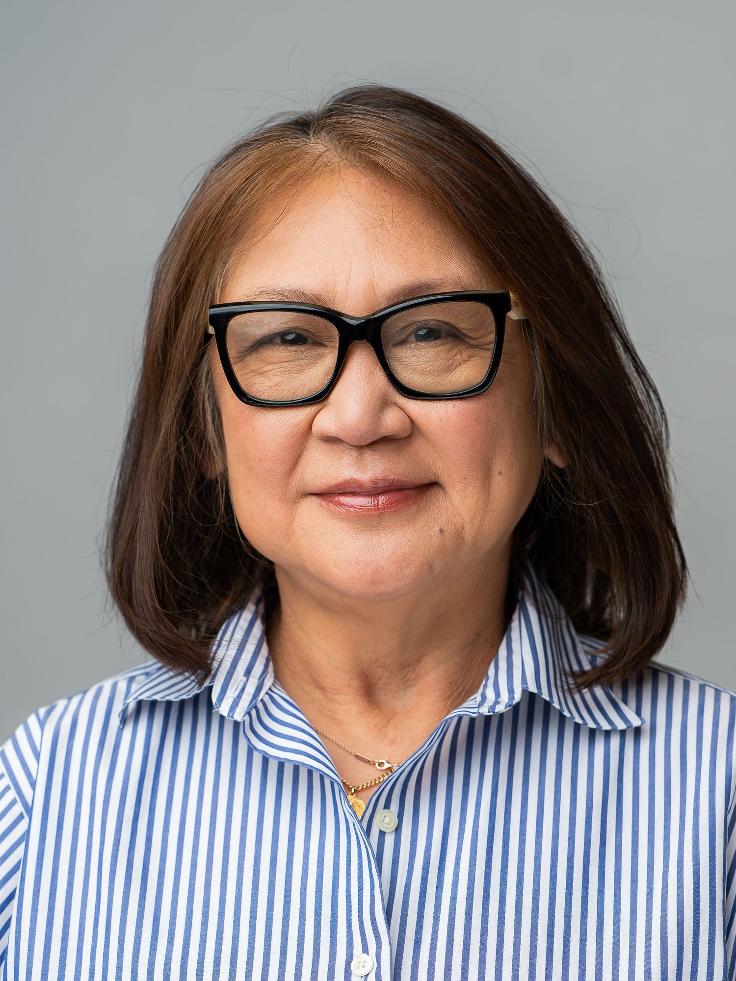 A middle-aged woman with black glasses and brown hair wearing a blue striped shirt poses for a professional headshot.