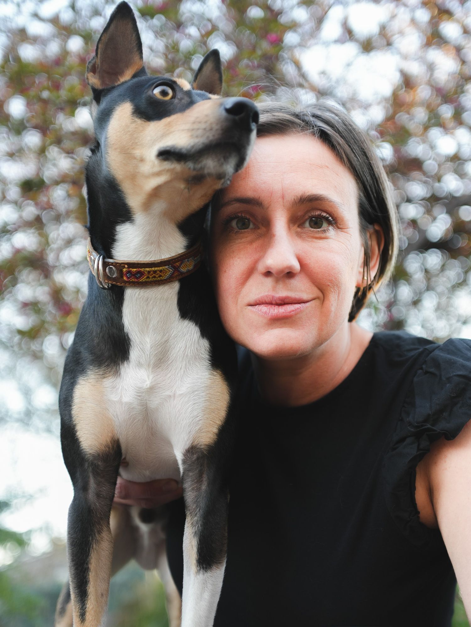 Woman taking a selfie with a black and white dog resting its head on her shoulder outdoors.