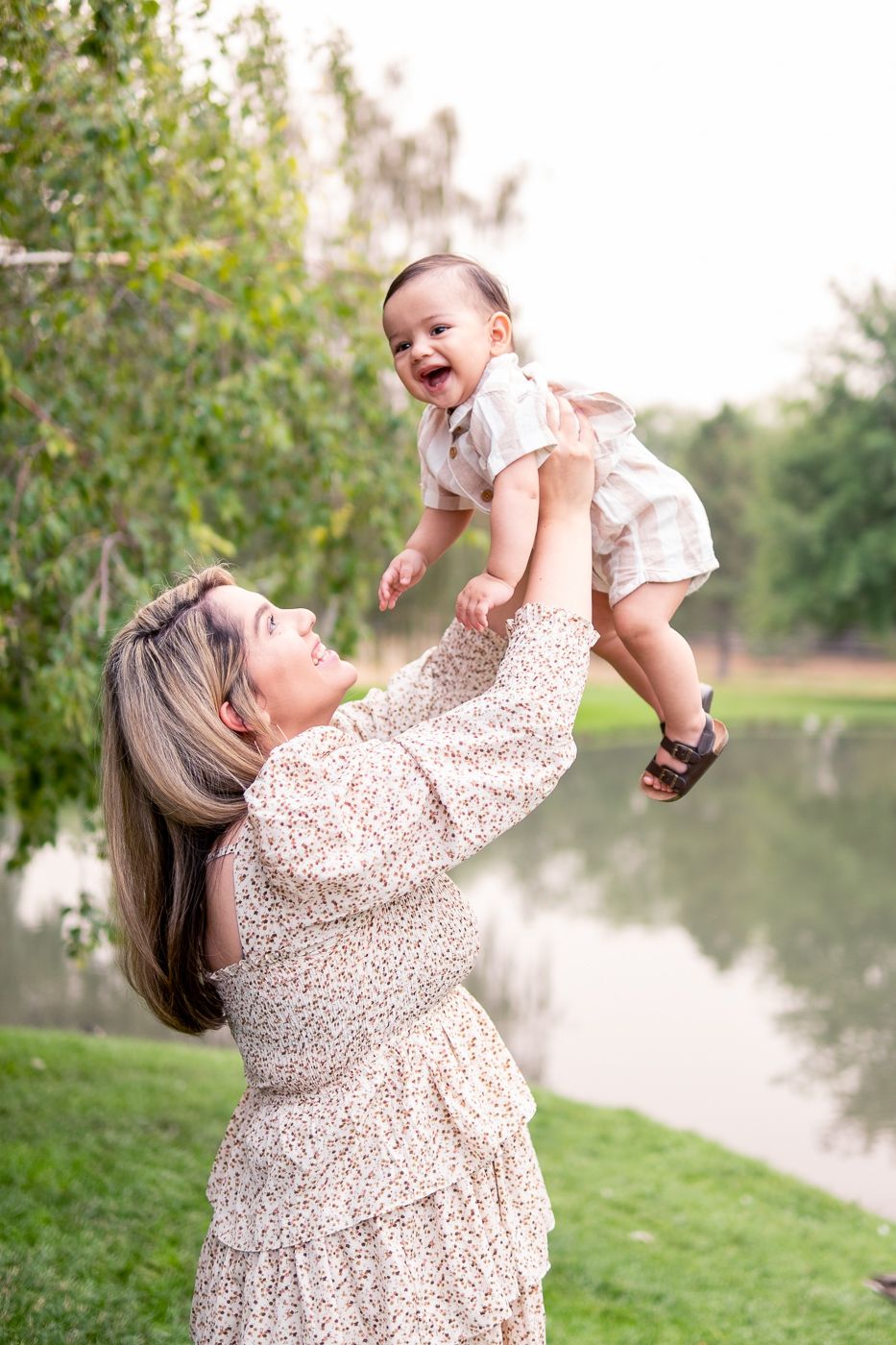 Woman in floral dress lifts laughing baby by a lake, surrounded by greenery on a sunny day.