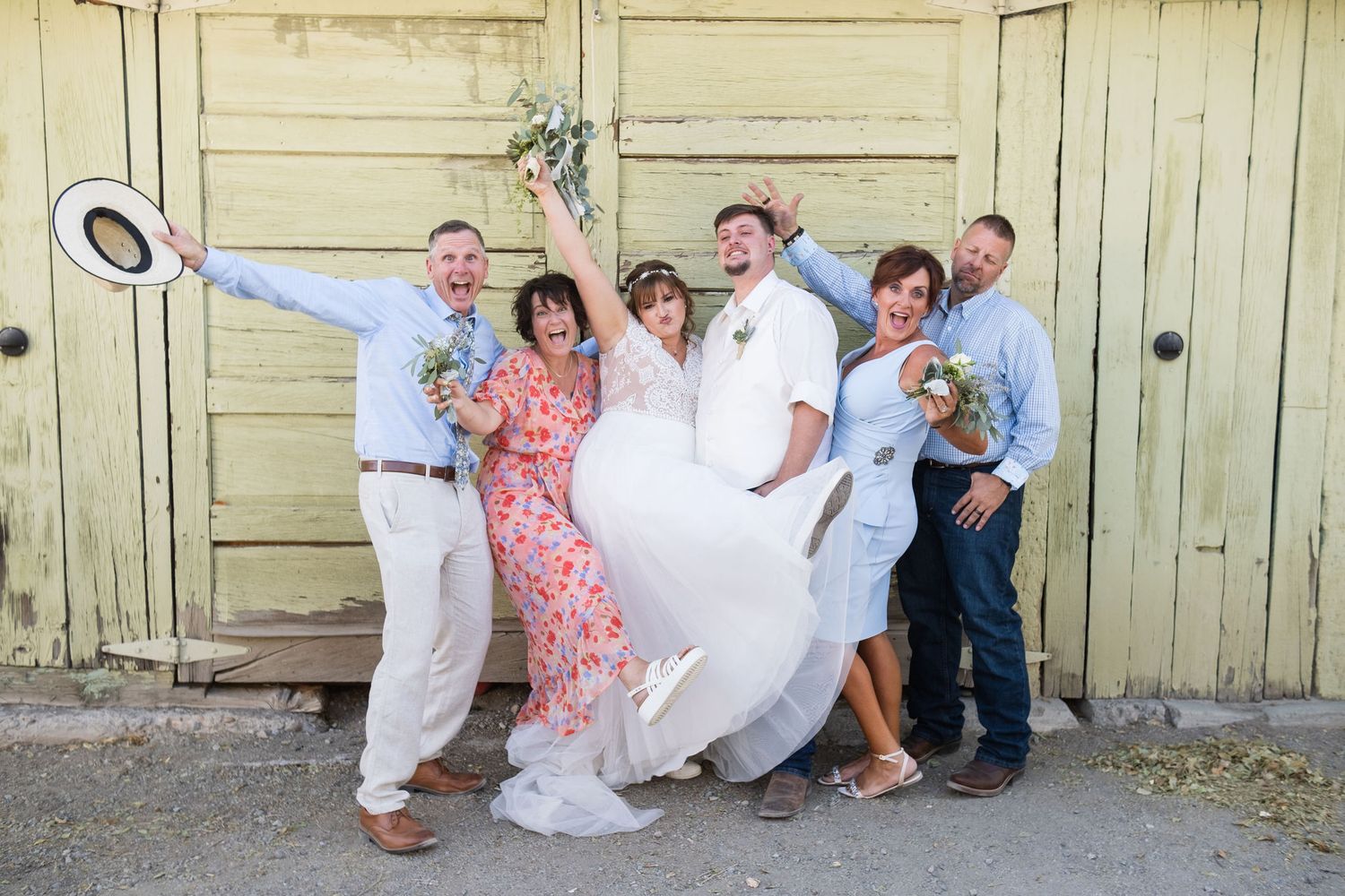 Joyful wedding party celebrating in front of rustic yellow doors, bride in white gown lifted by groom and family.