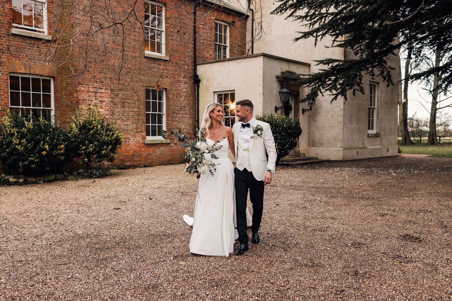 A couple in wedding attire walk together on a gravel path outside a historic brick mansion on their special day.