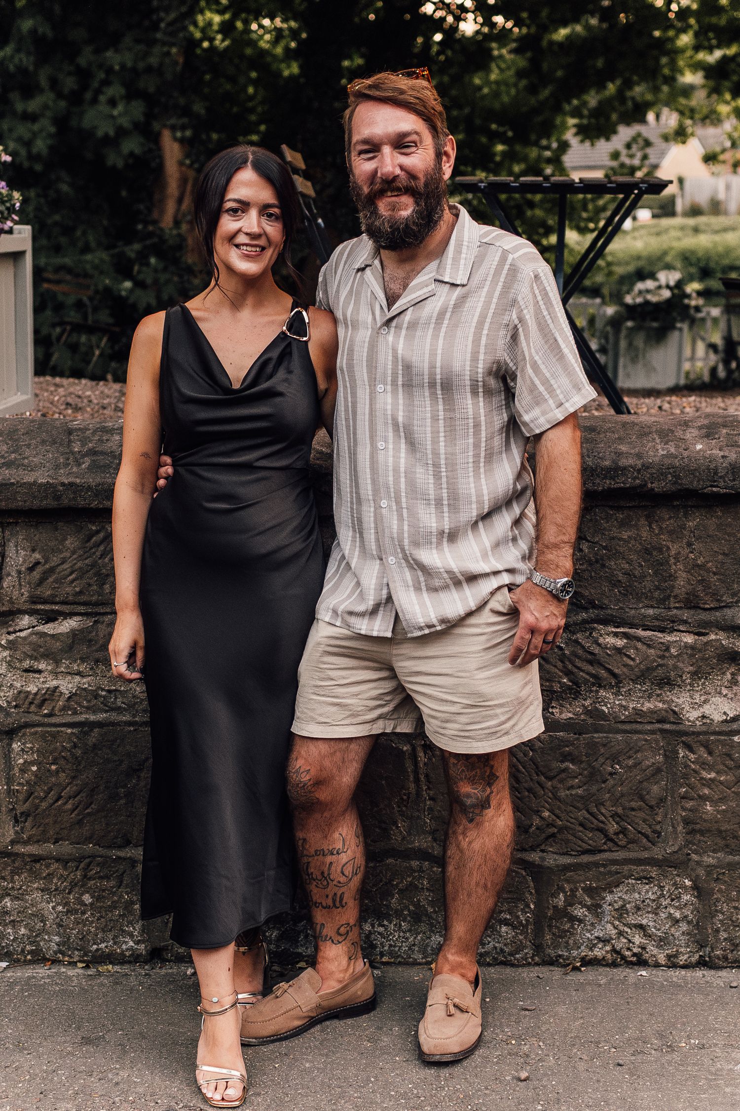 A smiling couple poses outdoors, woman in black satin dress and man in striped shirt with khaki shorts and leg tattoo.