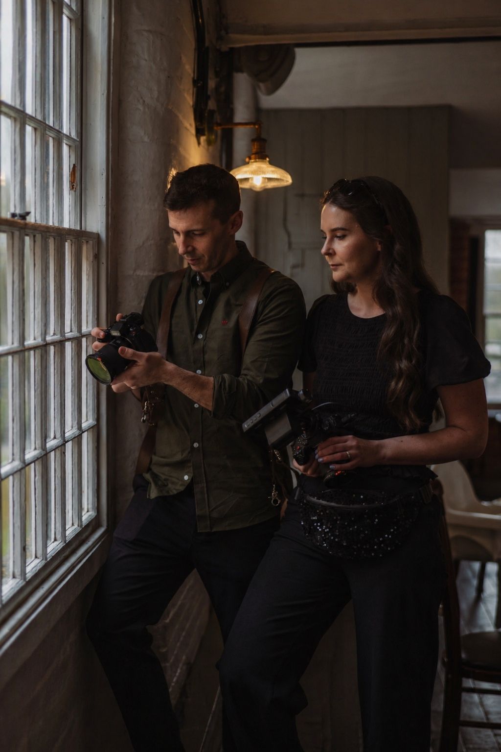 A man and woman stand by a window in dim light, reviewing photos on a camera together indoors.