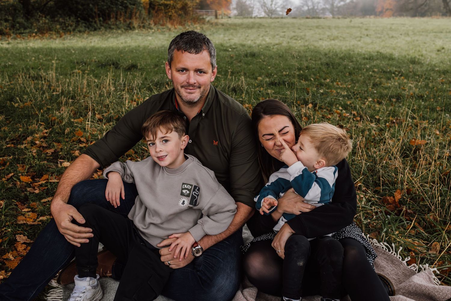 A family group sits together in a grassy autumn field during an outdoor photo session.