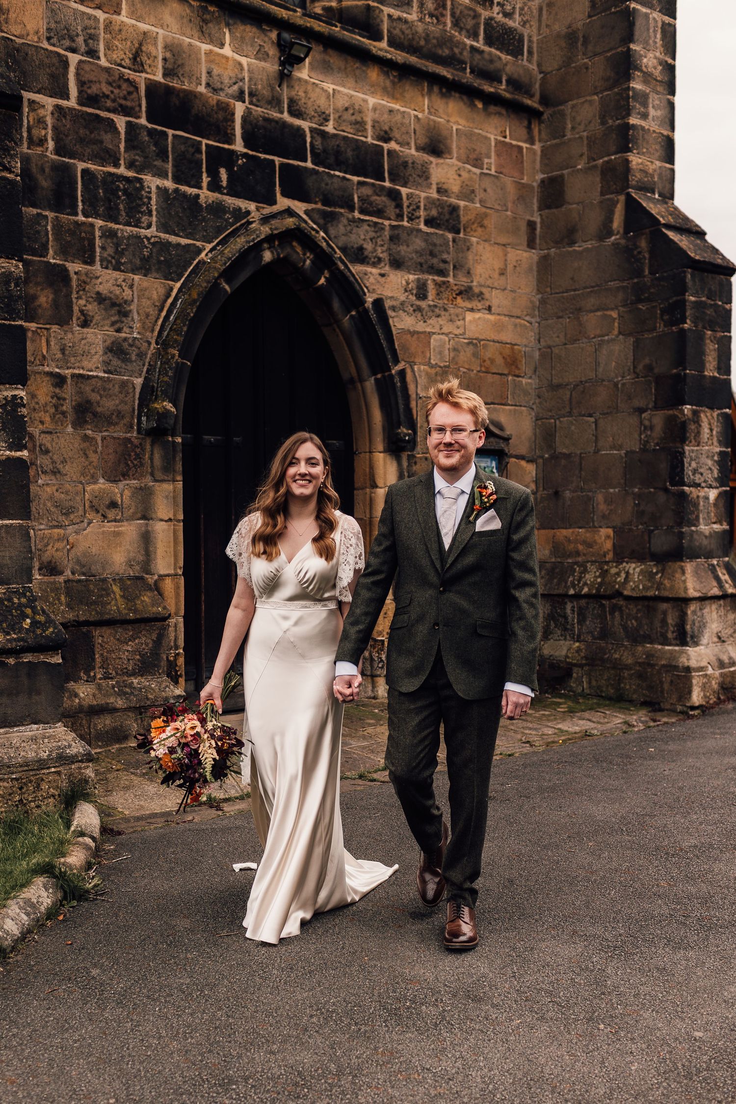 A couple in wedding attire walks and laughs together along a stone church exterior wall.
