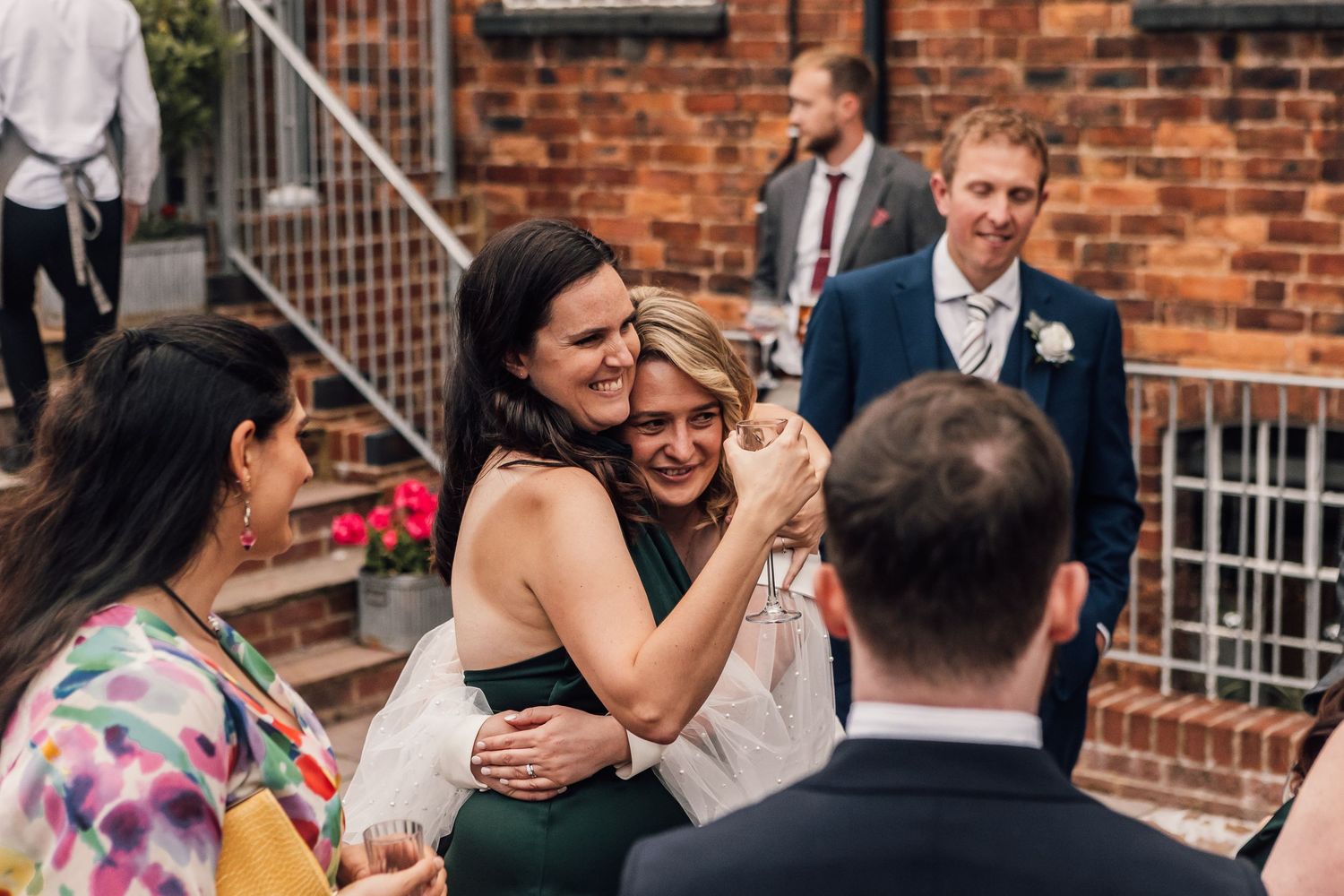Wedding guests embrace and share joyful moments during an outdoor reception at a rustic brick venue.