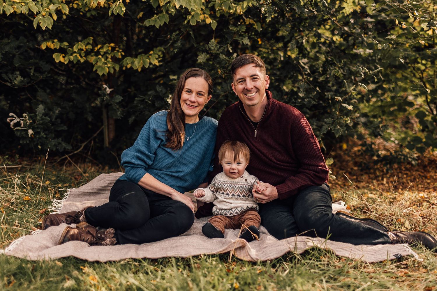 Family sitting together on a blanket outdoors during autumn with fallen leaves and greenery in the background.