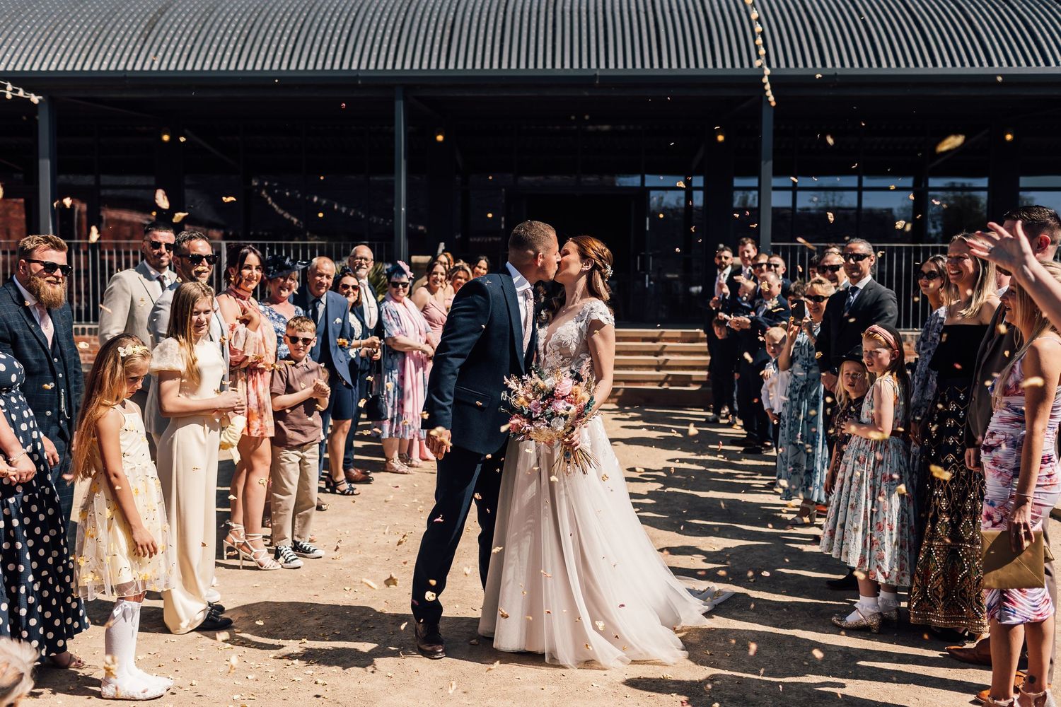 Bride and groom share a kiss while guests throw confetti during their outdoor wedding ceremony exit.