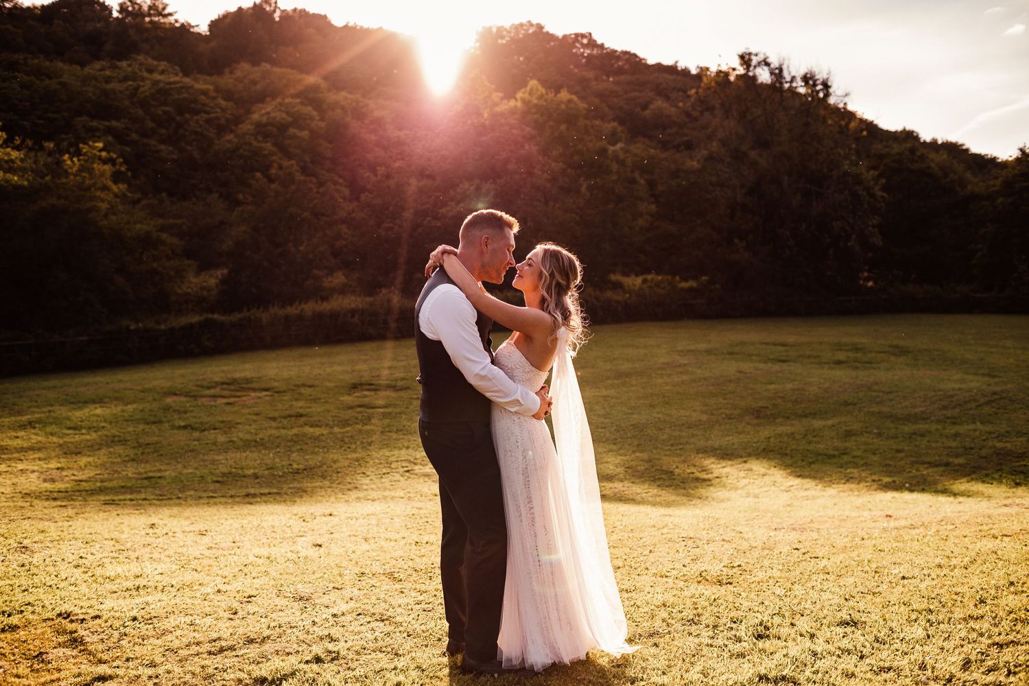 A newlywed couple embraces in a golden field at sunset with trees silhouetted in the background.