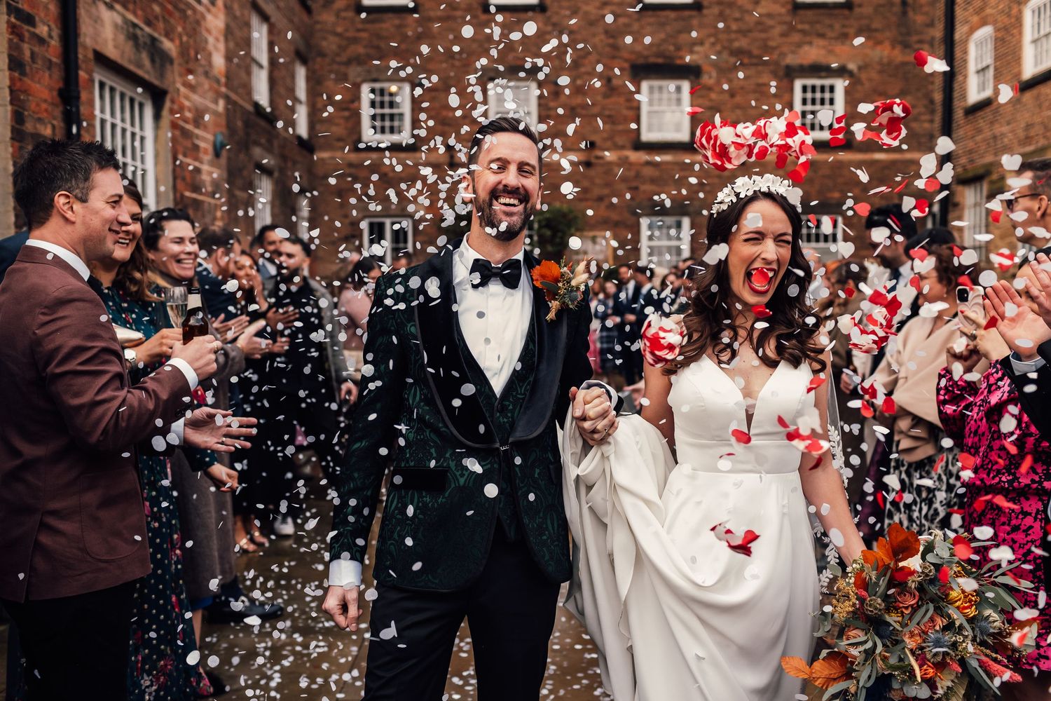 A joyful wedding couple exits their ceremony while guests shower them with confetti and rose petals at a brick venue.