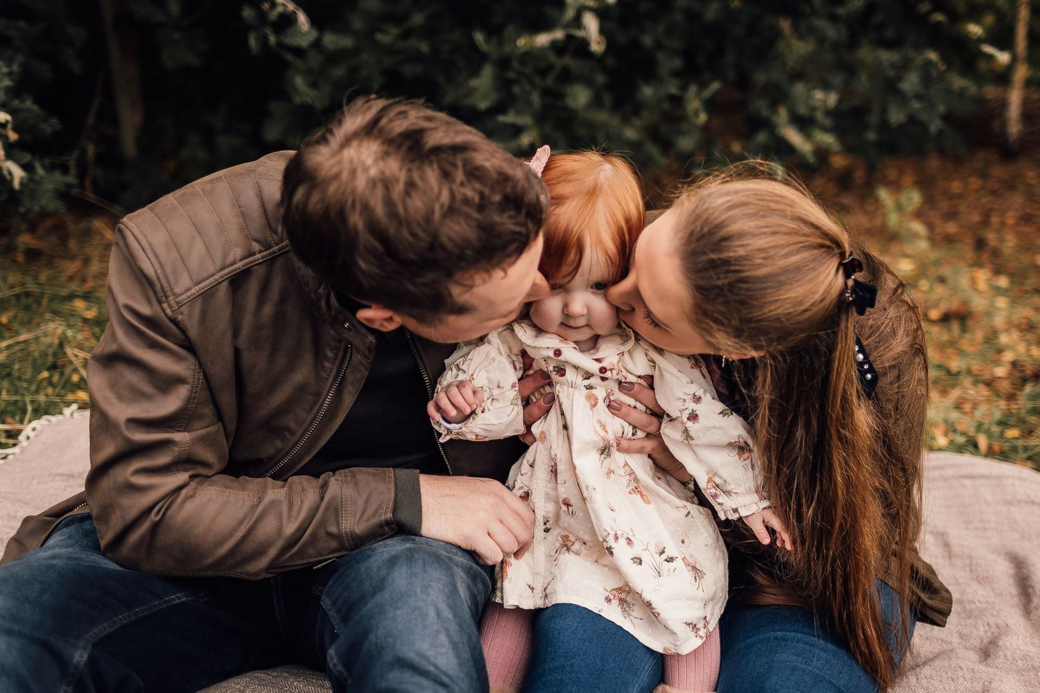 Loving family with small child sits together on blanket outdoors while sharing playful moment in autumn setting.