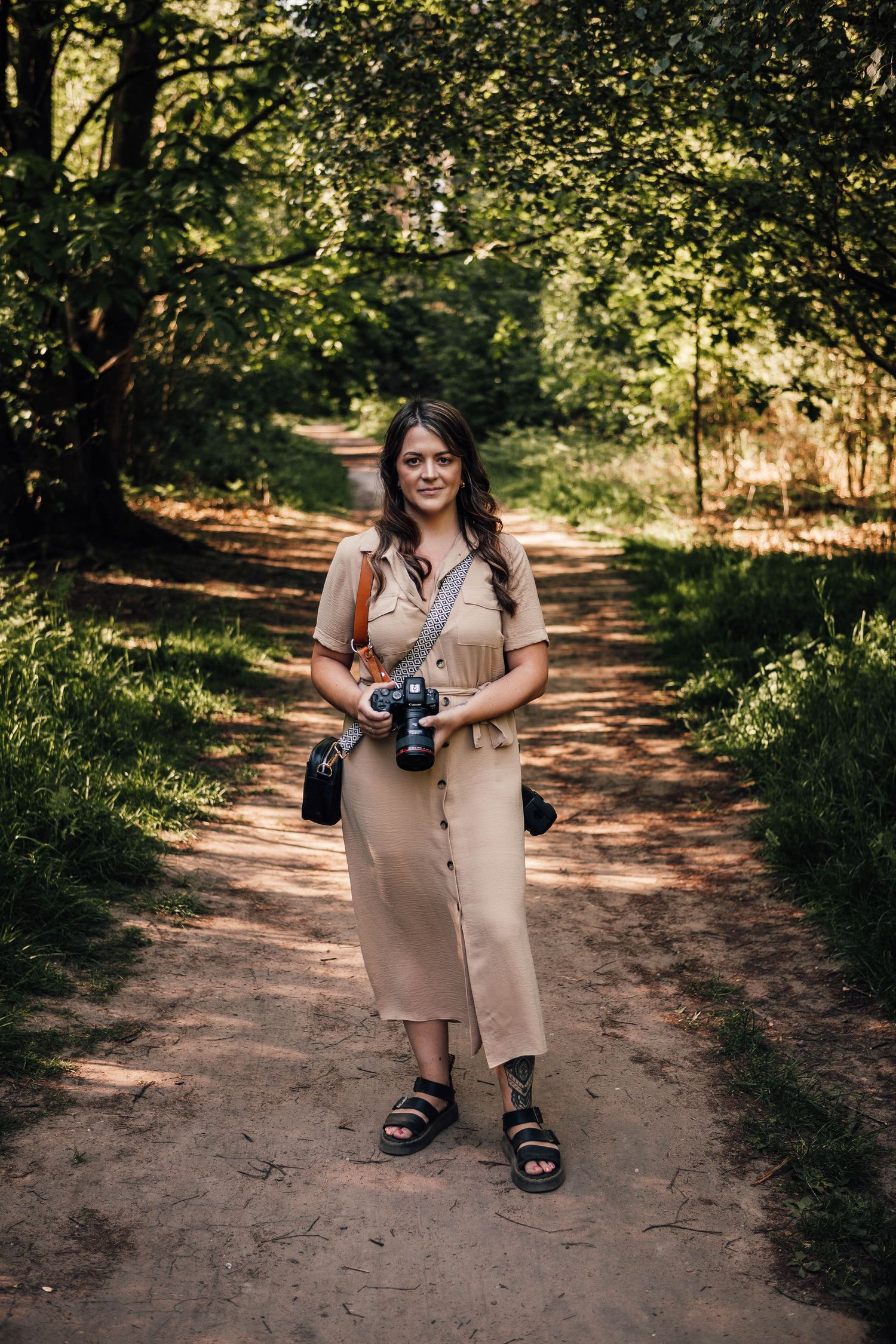 A photographer in a beige button-down dress and sandals stands on a sunlit forest path holding a camera.