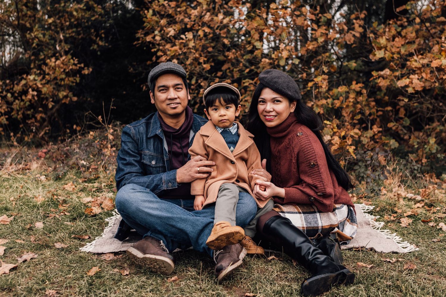 A family sits together on a blanket outdoors during autumn with orange and brown foliage in the background.