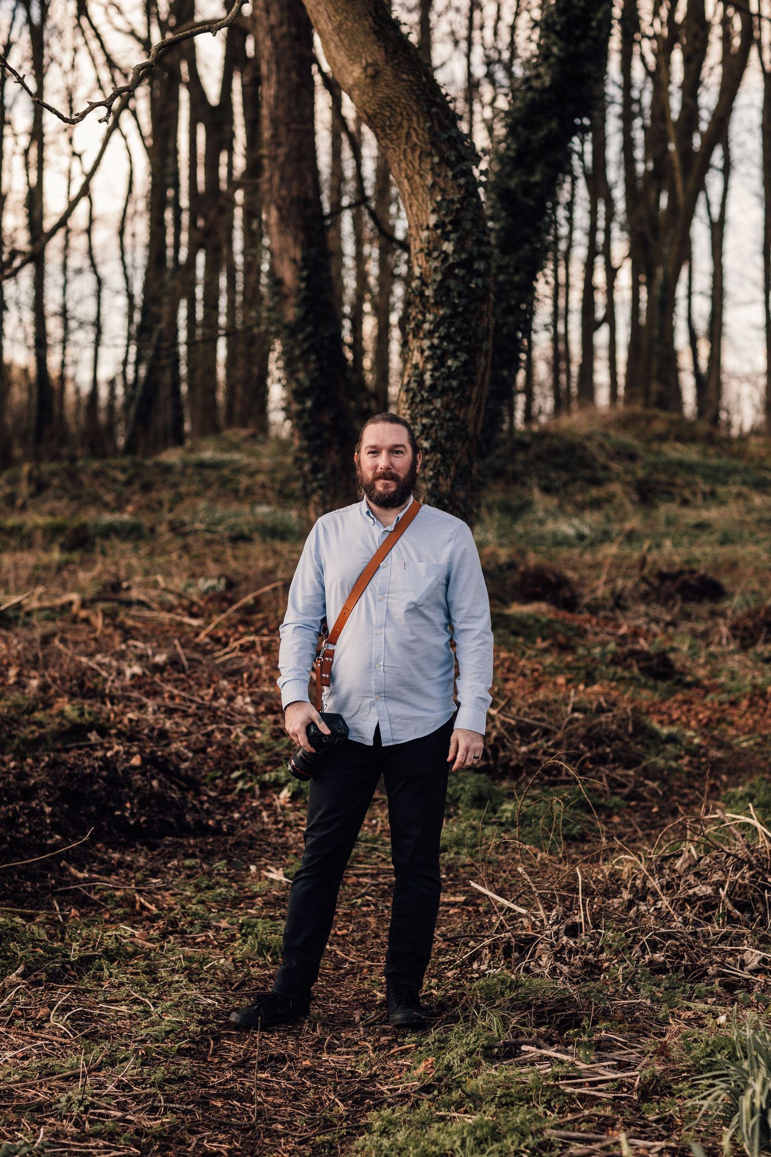 A person stands in a wooded forest setting wearing a light blue button-up shirt and dark pants with a leather bag.