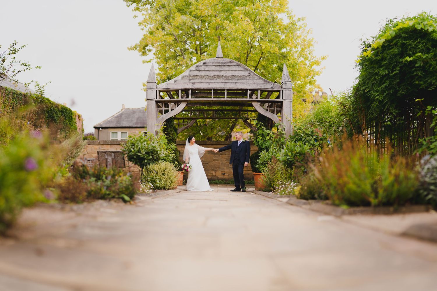 Two figures walk hand in hand on a garden path with lush greenery and architectural elements in the background.