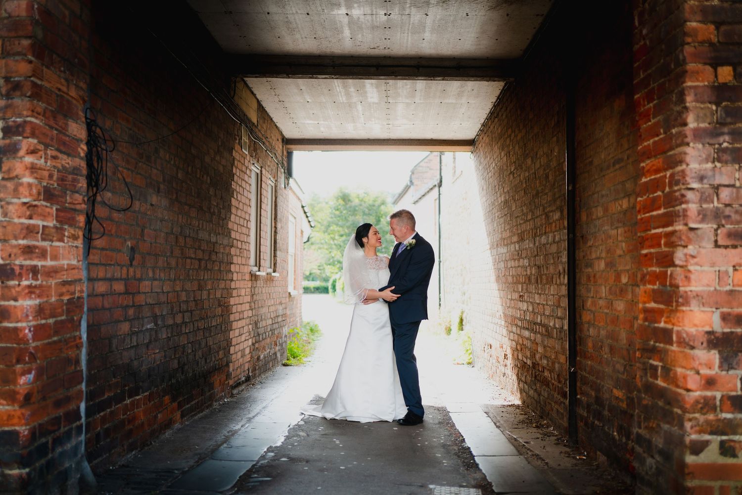 Silhouettes stand in romantic brick tunnel walkway with natural light streaming through both ends in wedding photo series.