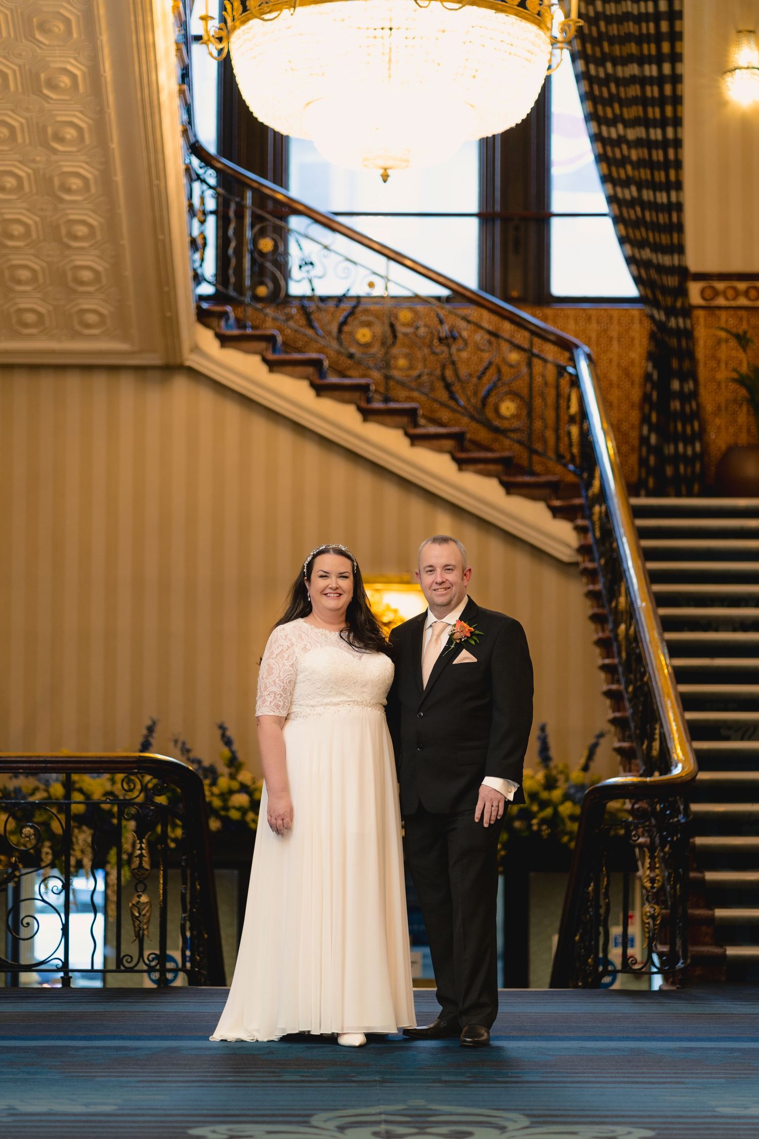 A couple in formal wedding attire poses elegantly on elegant hotel stairs with ornate railings and carpeting.