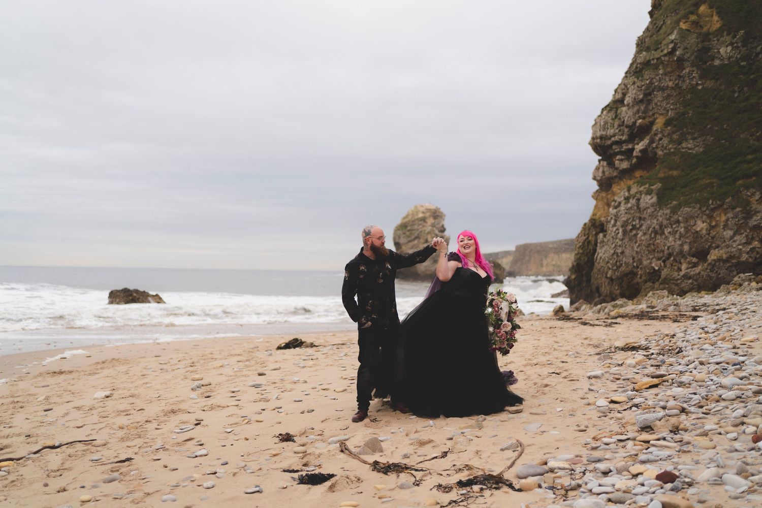 Two people in dark clothing sit on a sandy beach near rocky cliffs while looking out at the ocean waves.