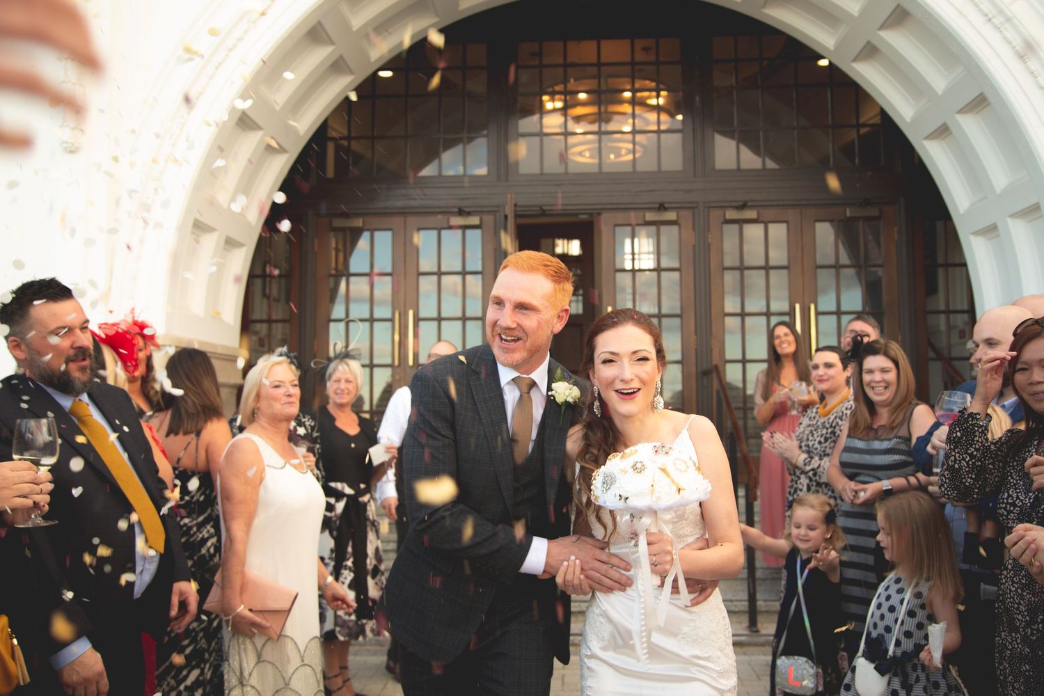 A group of wedding guests celebrate and dance together under elegant arched doorways at an indoor reception venue.