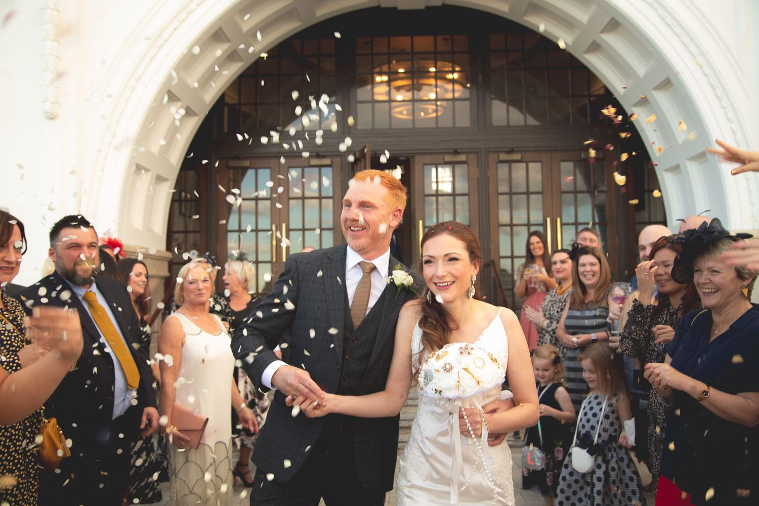 Couple exits venue under shower of confetti while guests celebrate.