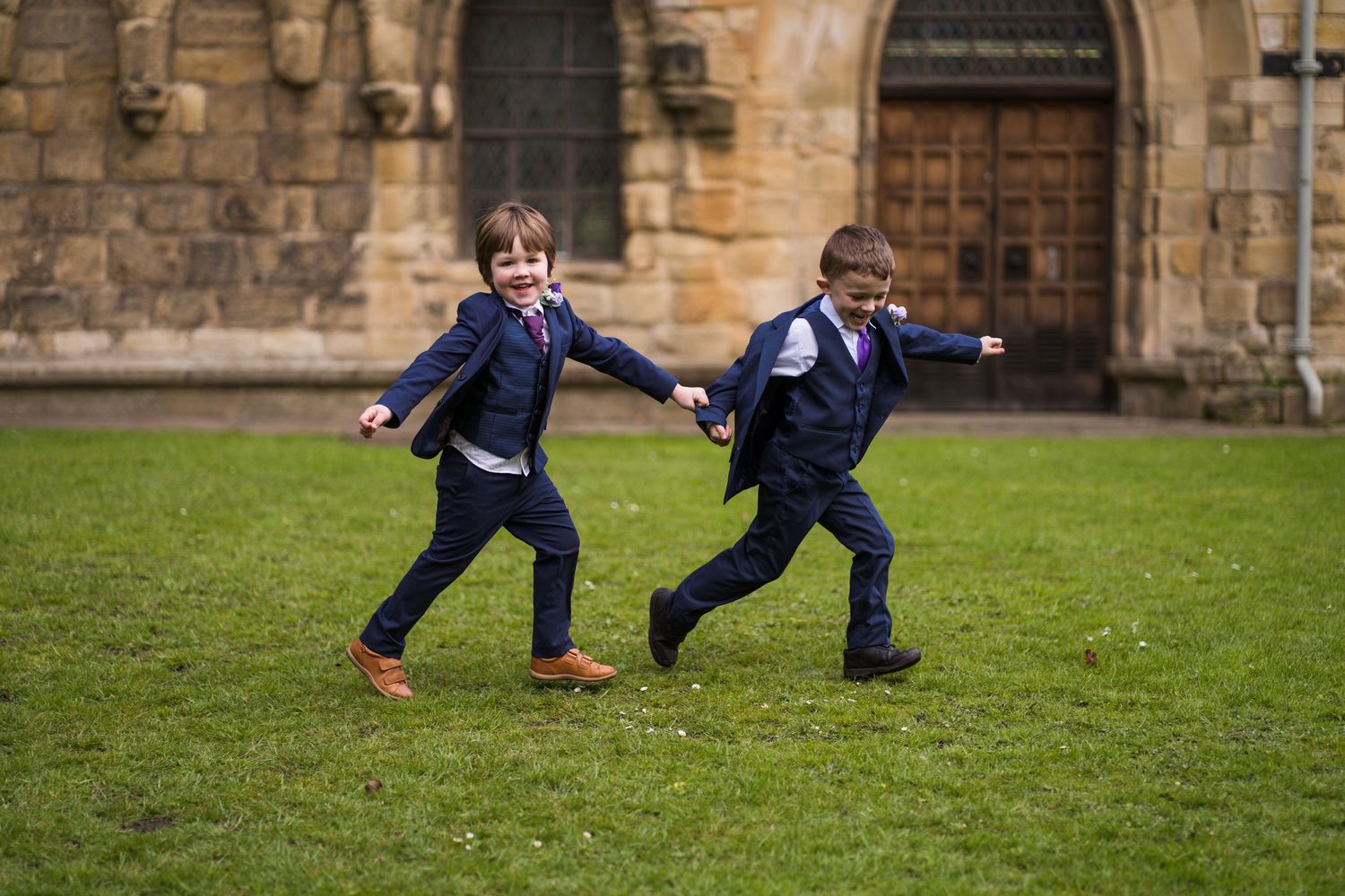 Two boys playfully pose while running on a green lawn near a historic stone building's exterior.