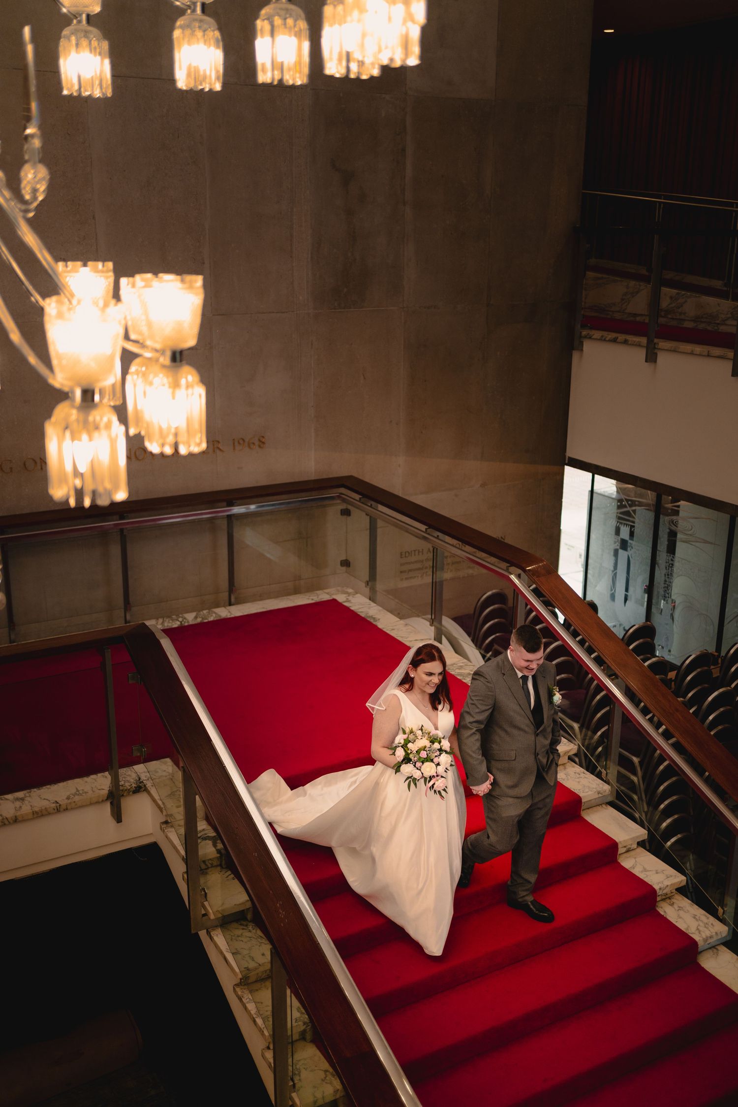 A couple descends a grand staircase with red carpet and vintage crystal chandeliers in an elegant venue.