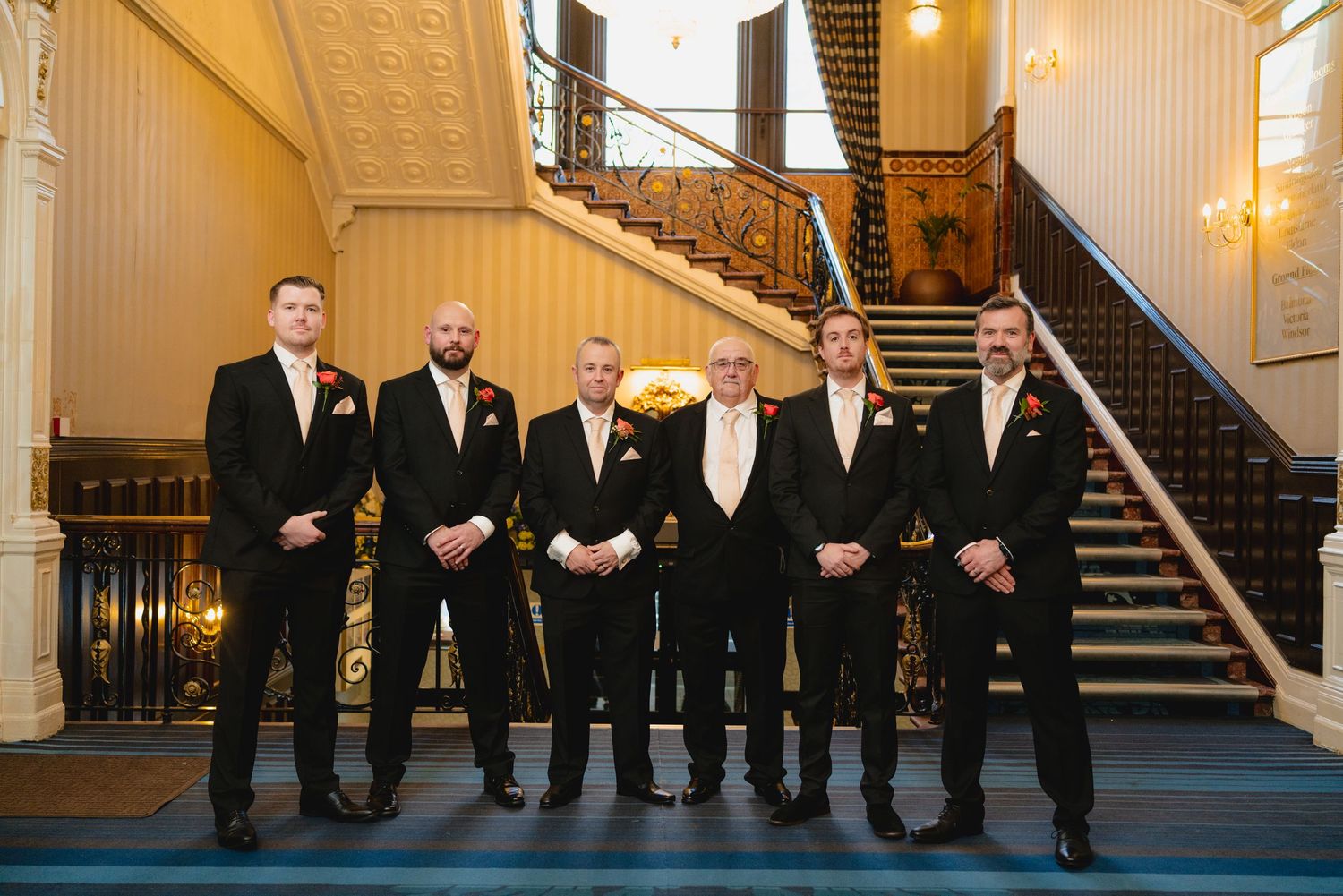 Groomsmen in black suits stand in elegant hallway with ornate staircase and warm lighting.