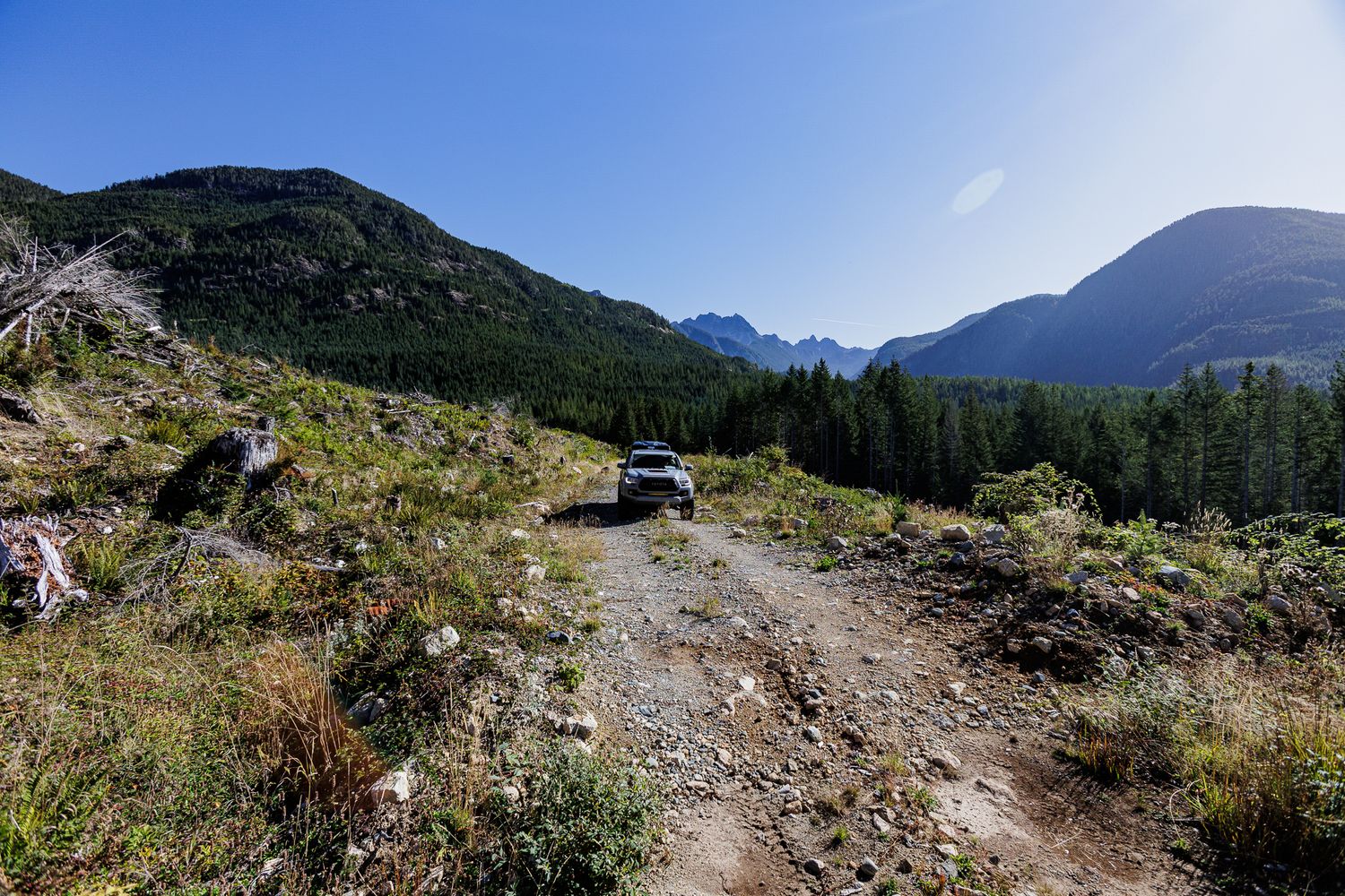 Dirt hiking trail winding through mountains with evergreen forest and turquoise camper van in distance under blue sky.