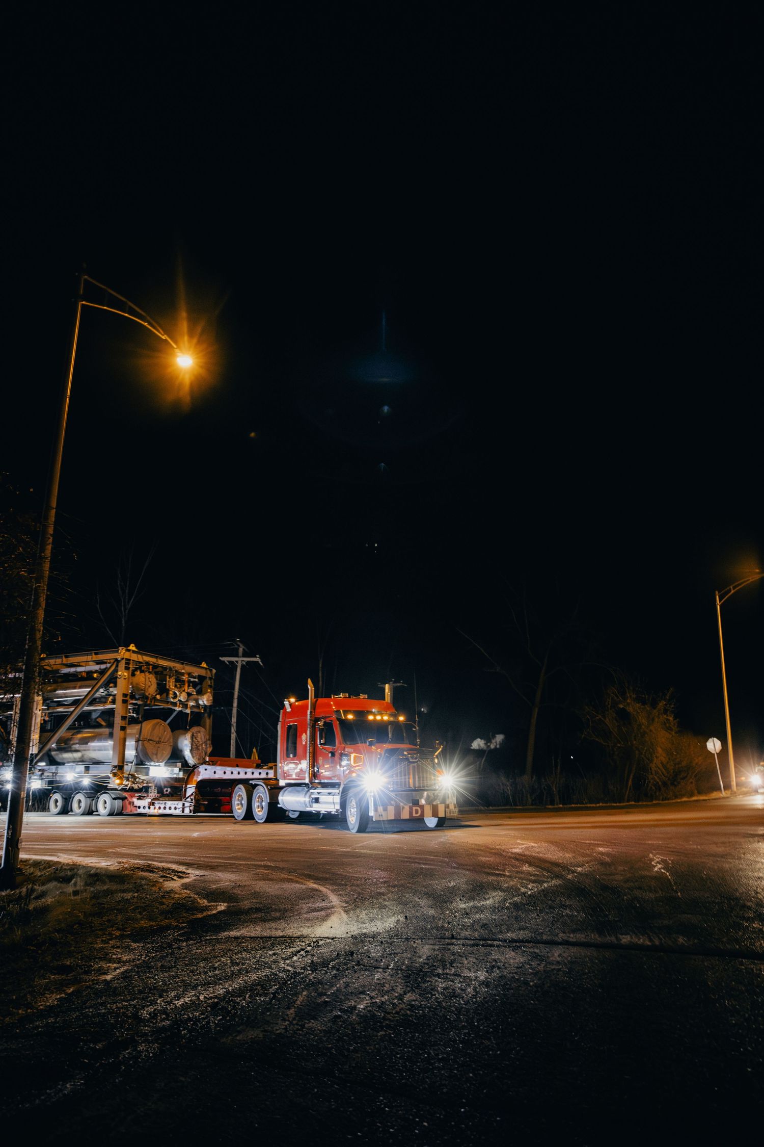 A semi truck illuminated by street lights makes its way down a dark road at night, with warm glowing lamps overhead.