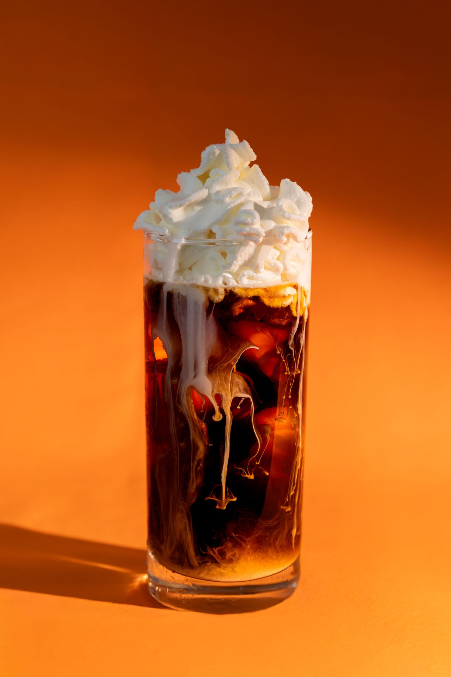 Glass of layered cold brew coffee with whipped cream topping against an orange background in dramatic lighting.