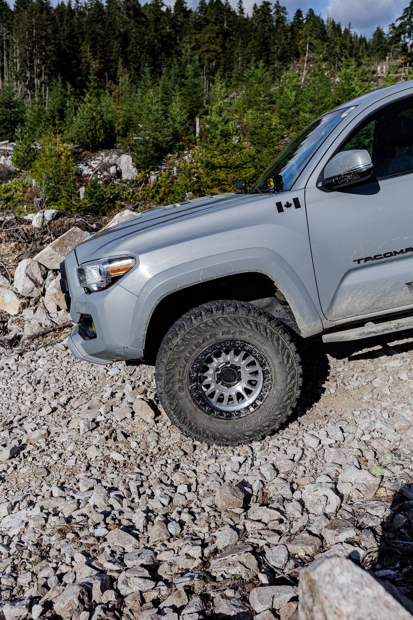 Silver Toyota Tacoma with off-road tires navigates rocky terrain on mountain trail with evergreen forest backdrop.