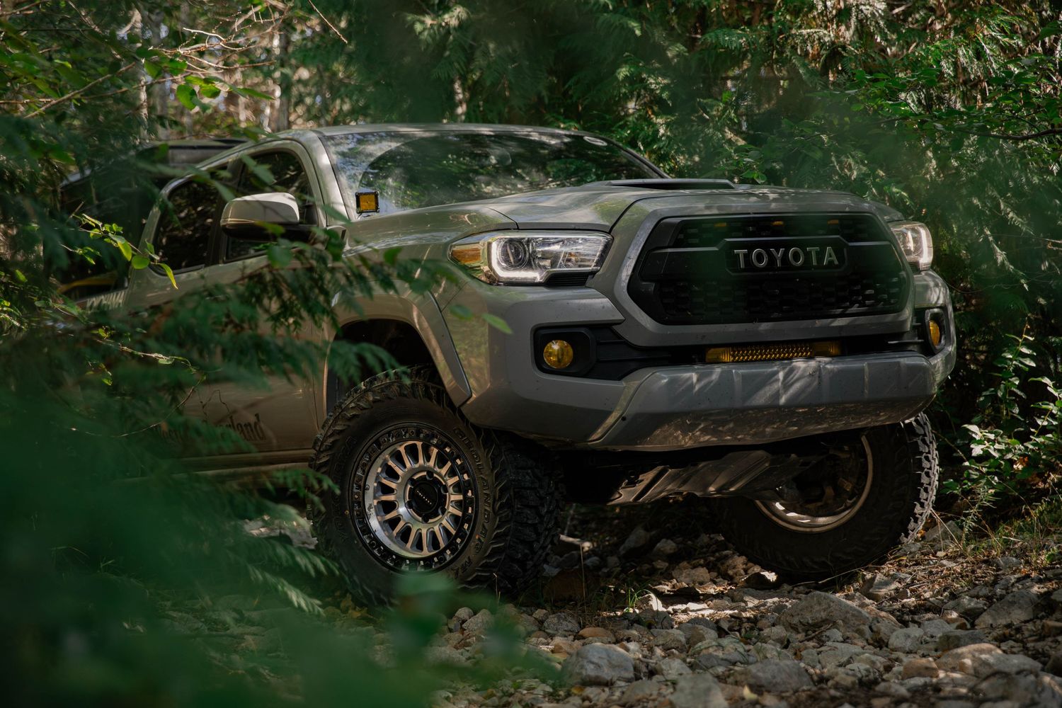 Green Toyota Tacoma truck with off-road modifications driving through rugged forest terrain.