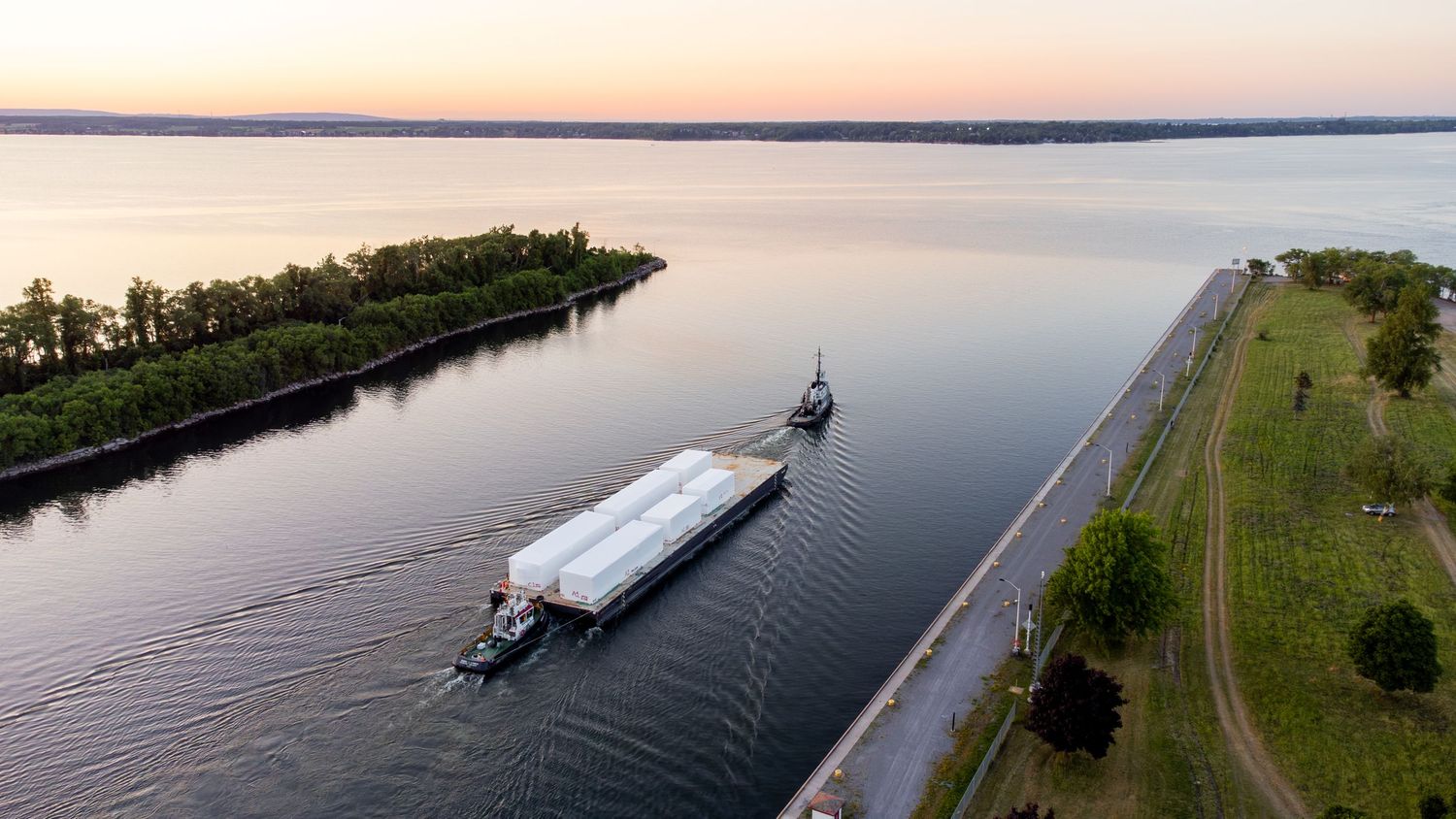 Aerial panoramic view of boats cruising along a scenic river channel surrounded by green shorelines during sunset.